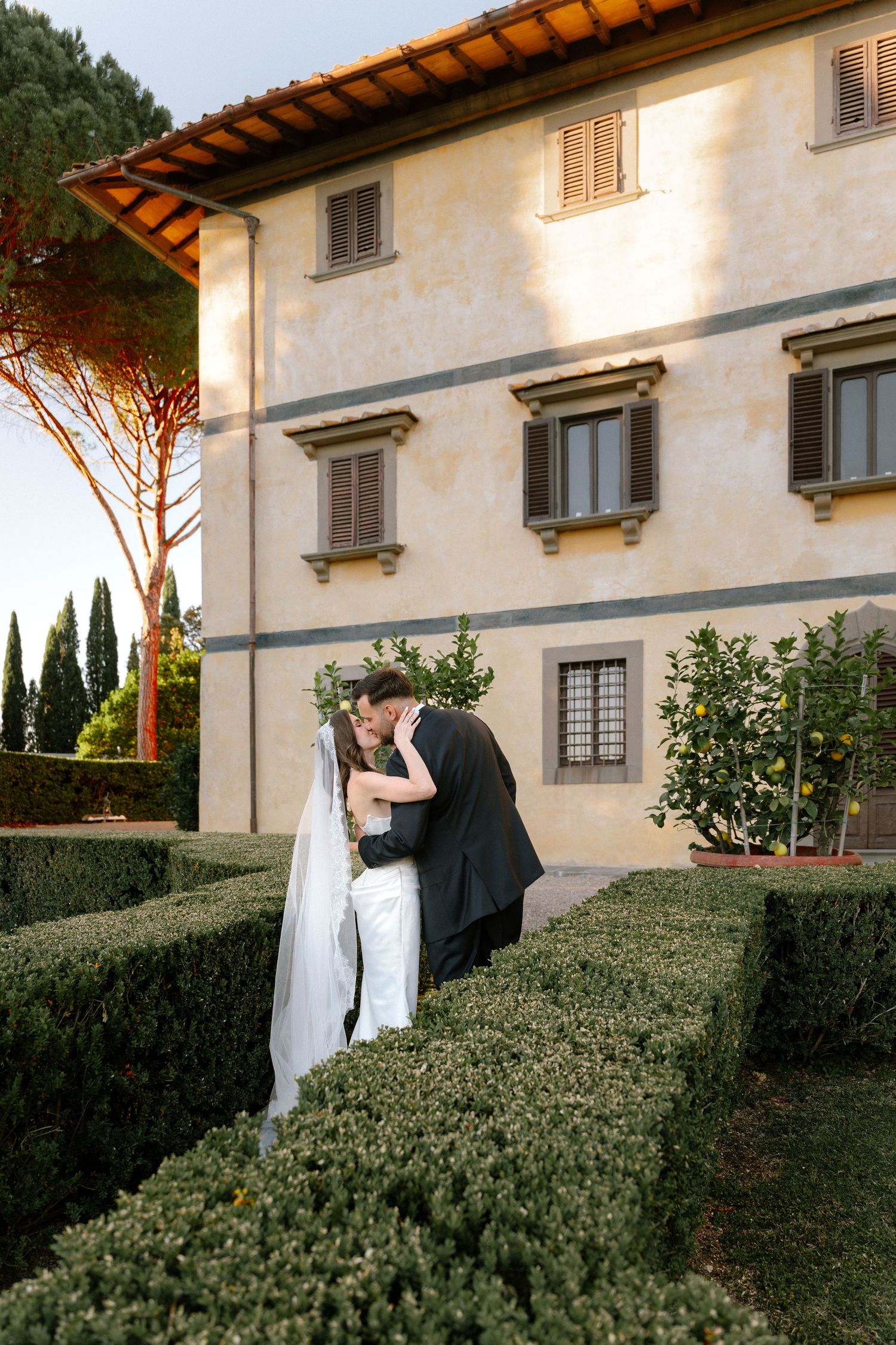 Bride and groom share a romantic kiss in a manicured garden beside a stunning Italian villa on their wedding day.