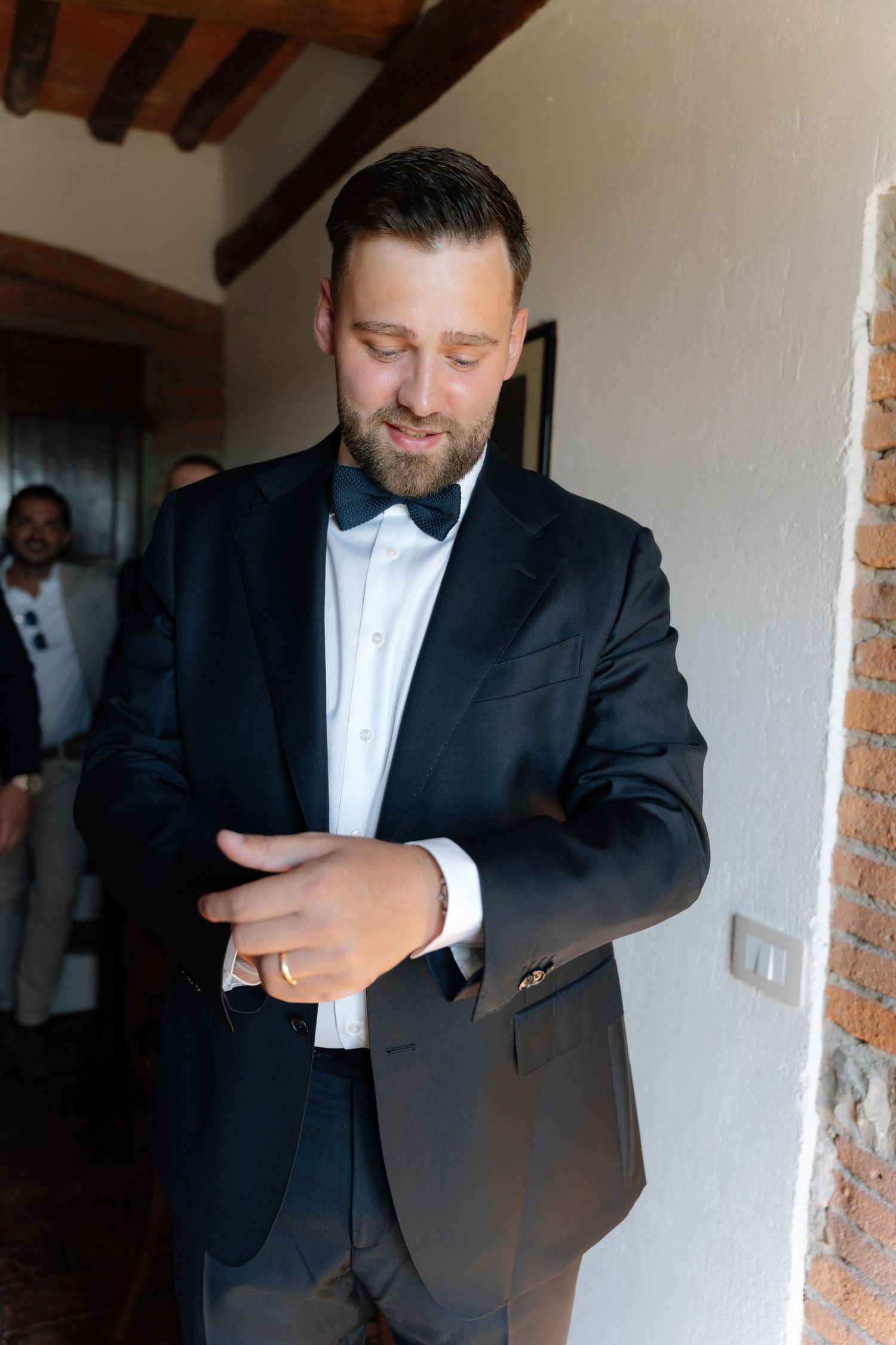 Groom in black tuxedo with bow tie adjusting cufflinks before wedding ceremony in rustic venue.