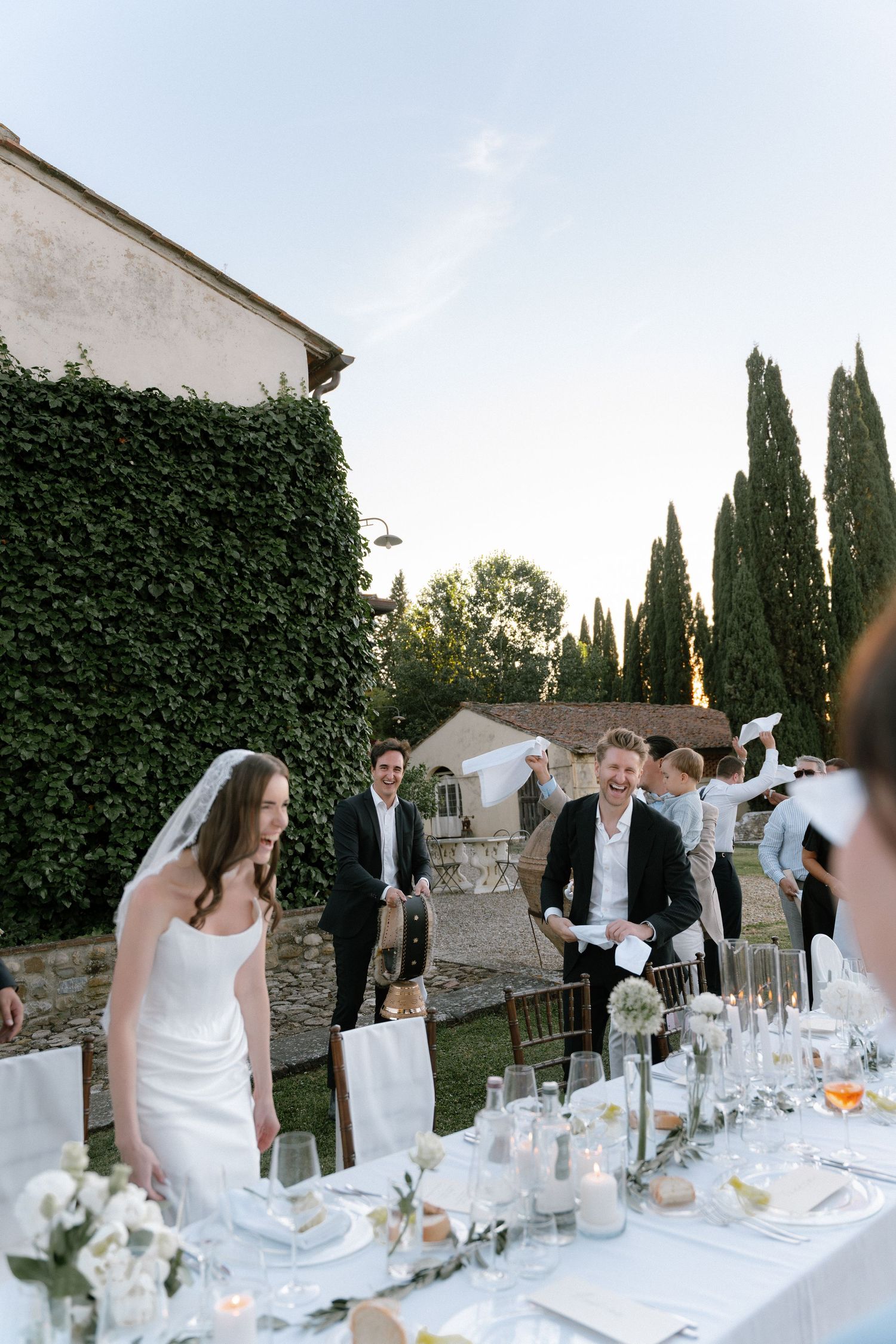 Bride and groom laugh at outdoor wedding reception table with white floral decor, cypress trees, and ivy-covered walls in Tuscany.