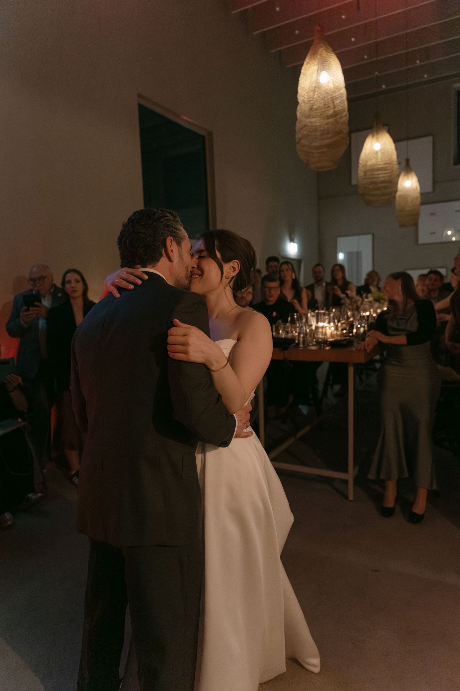 Bride and groom share first dance at wedding reception with guests watching and golden pendant lights above.