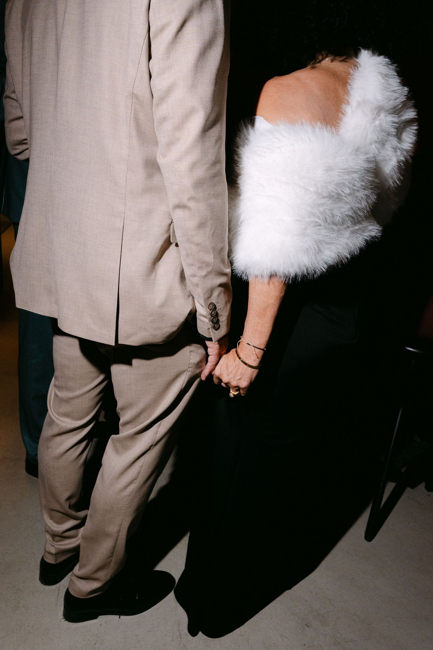 Couple holding hands, man in beige suit, woman in black dress with white fur stole, elegant formal attire.