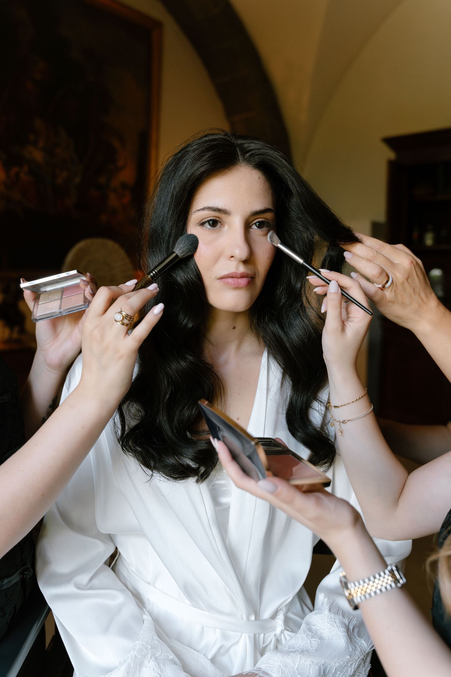 Bride getting makeup applied by two artists with brushes before her wedding ceremony.