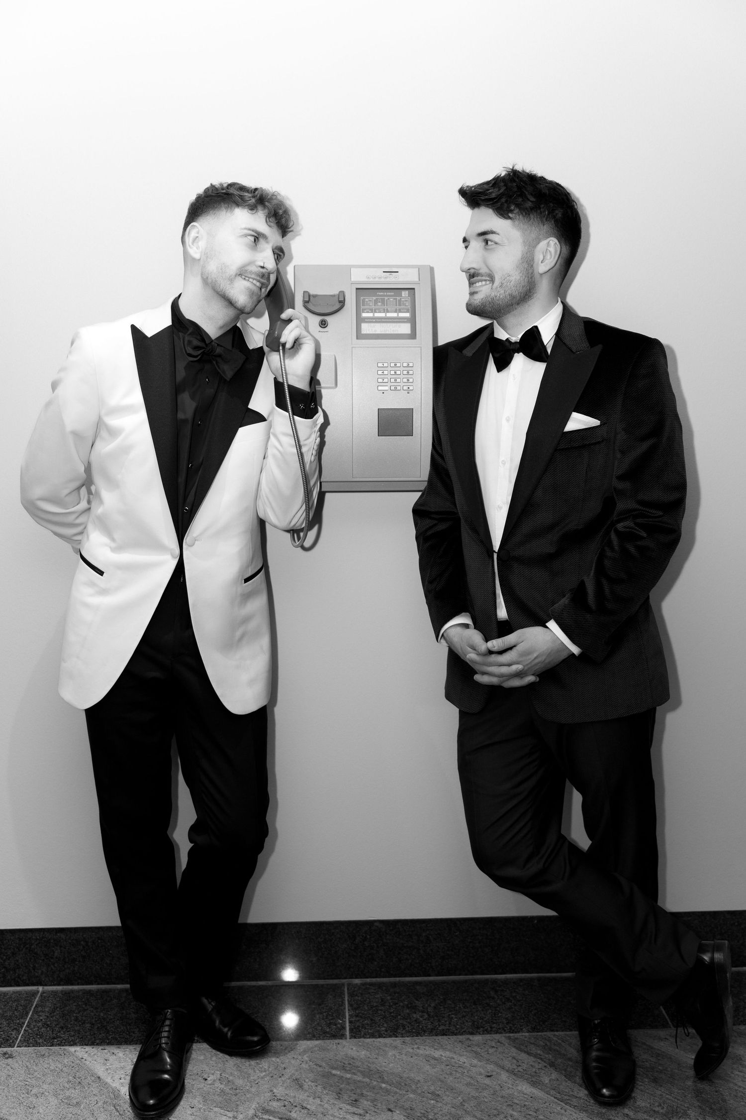 Two men in tuxedos pose by a payphone in a hallway, with one playfully using the phone while the other looks on.