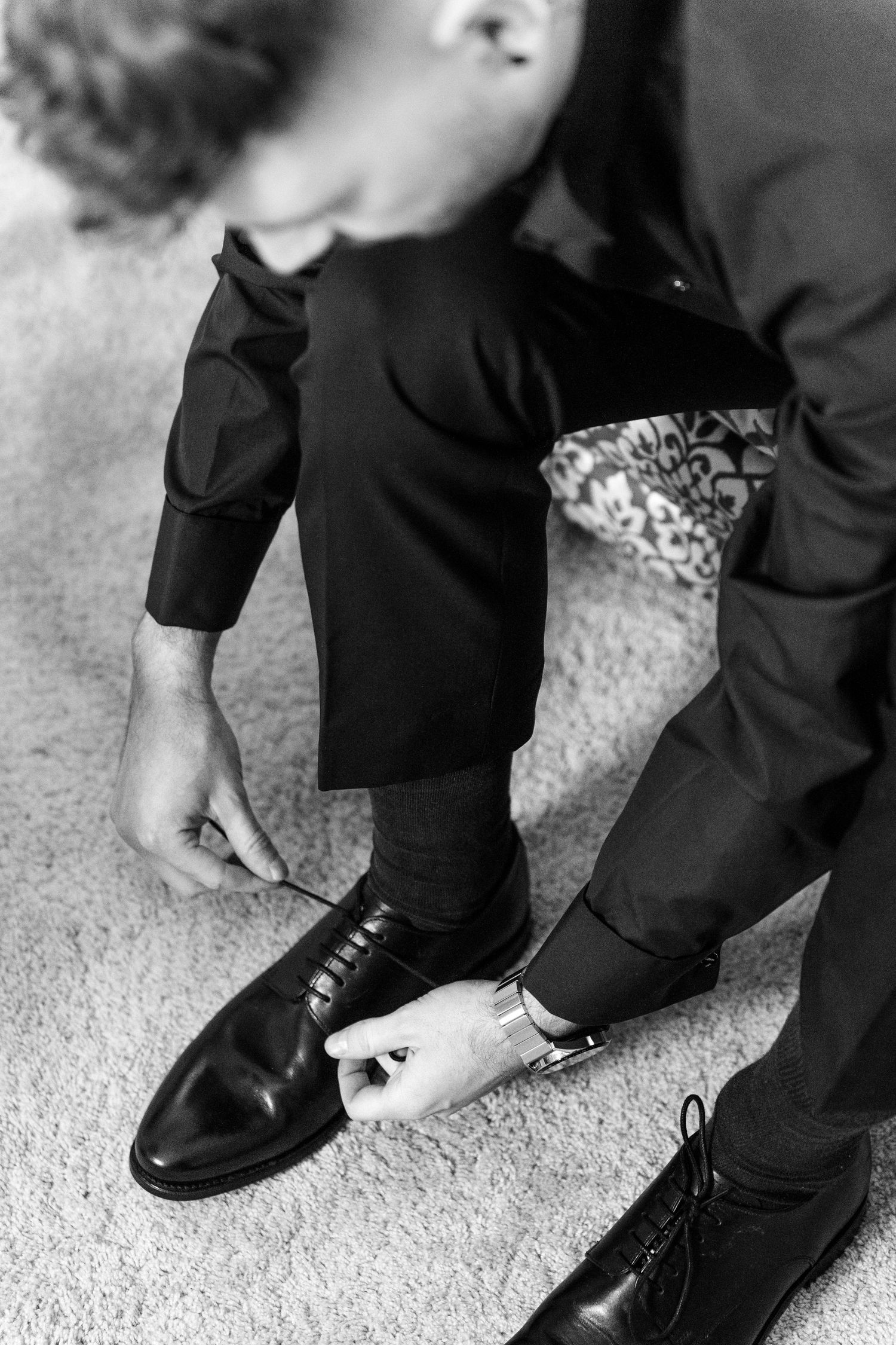 Black and white photo of hands lacing up polished dress shoes while getting ready on carpet.