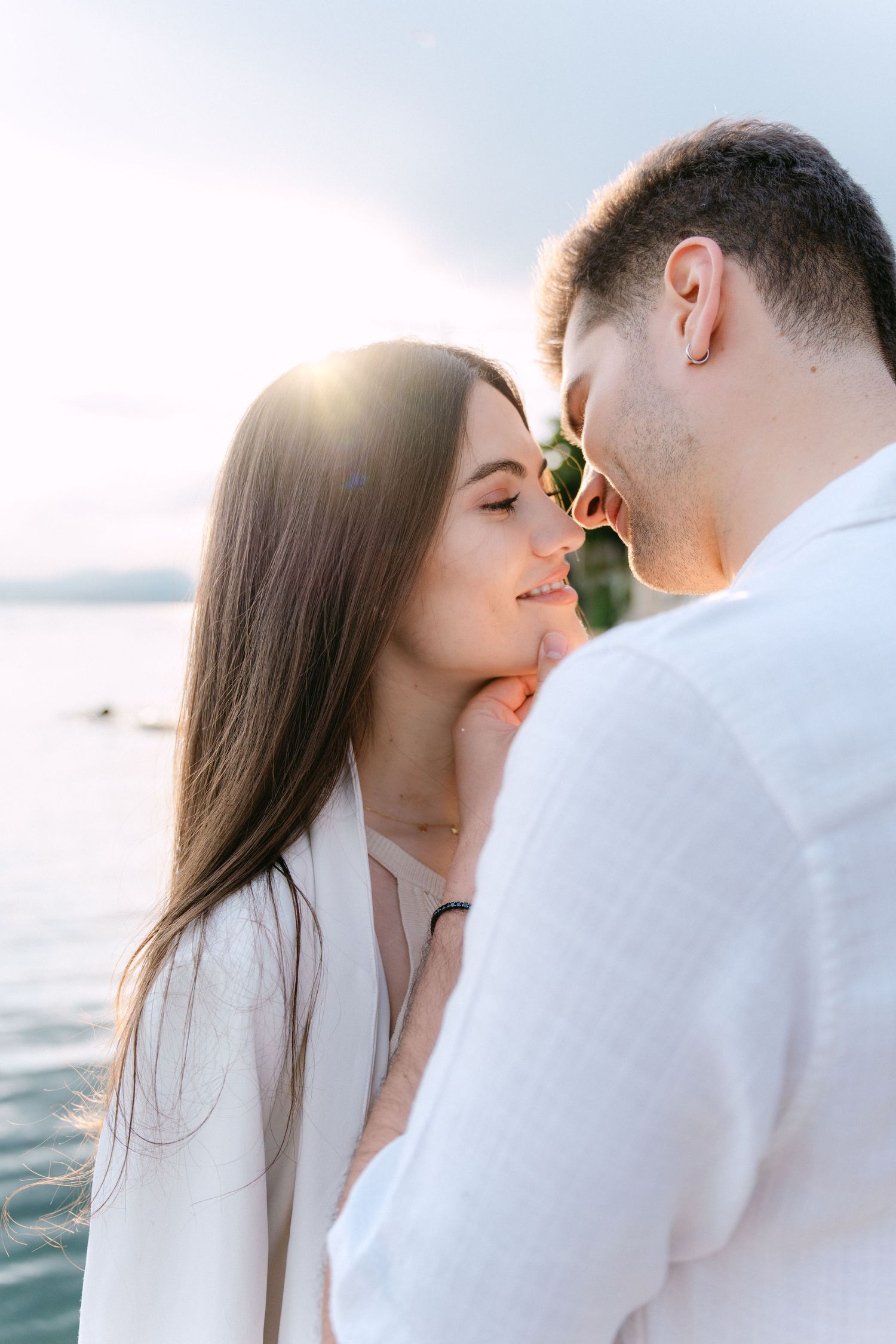 Romantic couple sharing intimate moments by the ocean at sunset in white outfits.