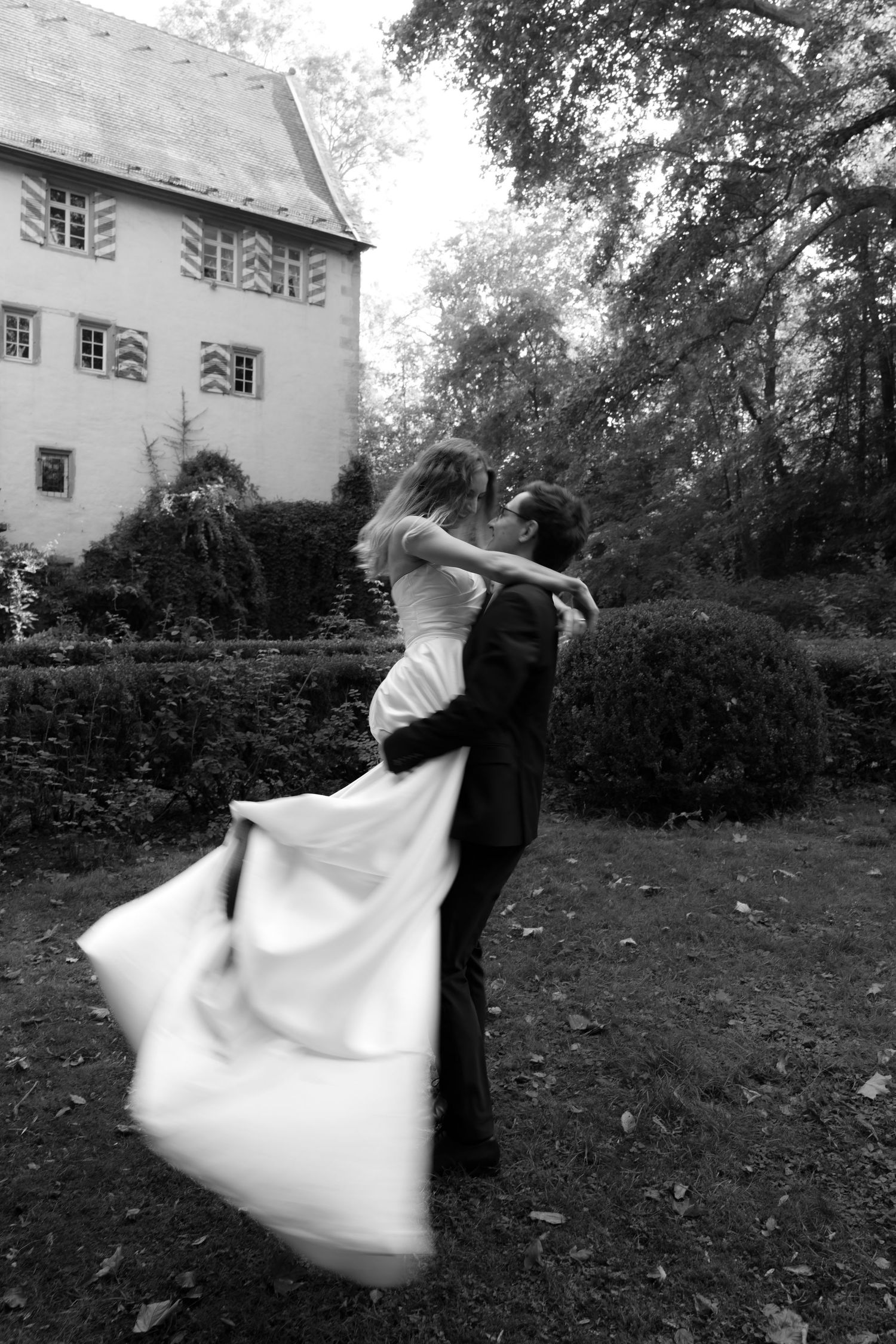 Black and white photo of a groom lifting and kissing his bride in a flowing gown on a lawn near a historic building and trimmed hedges.