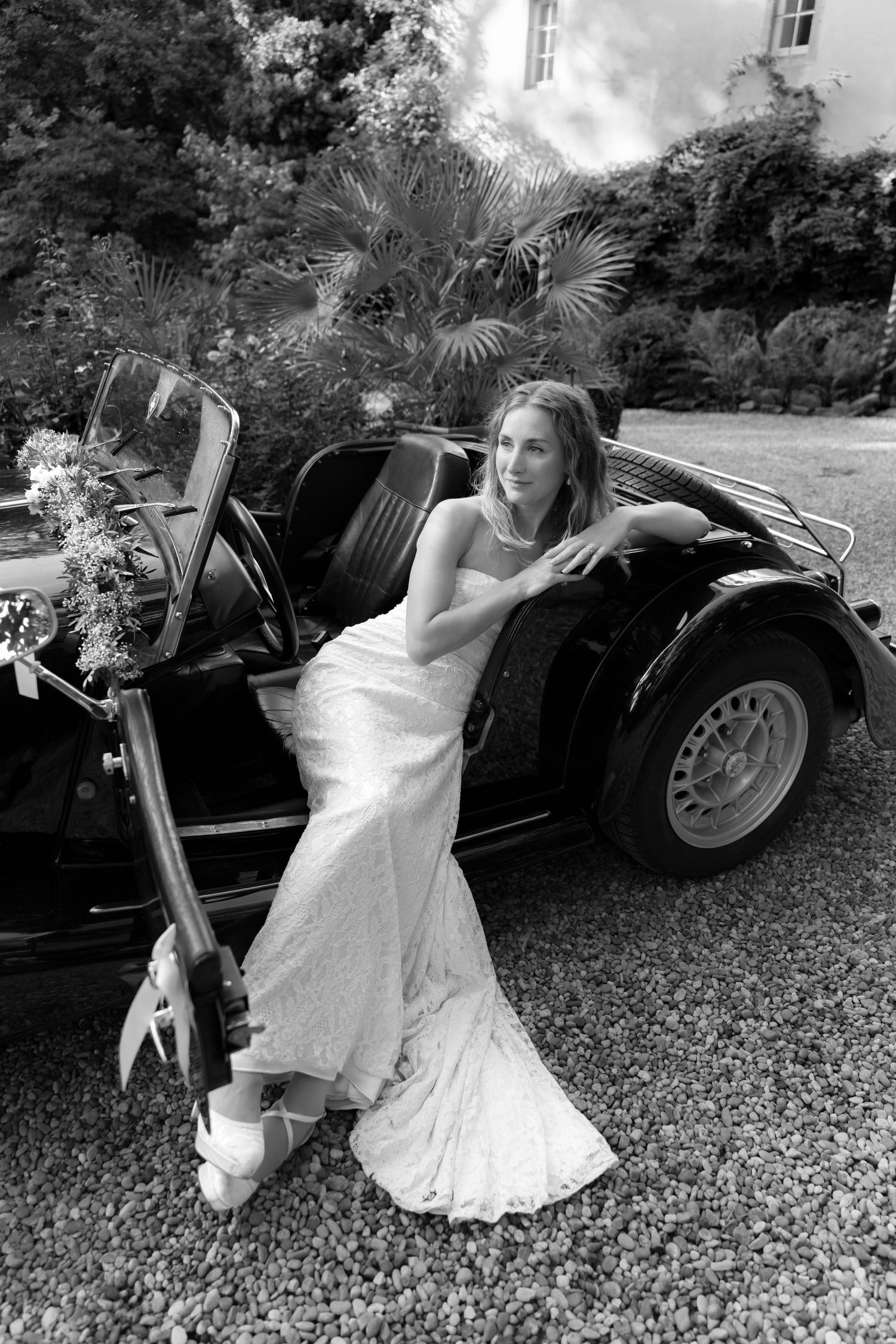 Bride in a strapless lace wedding gown sits sideways in a vintage black convertible car on a gravel drive, in black and white.