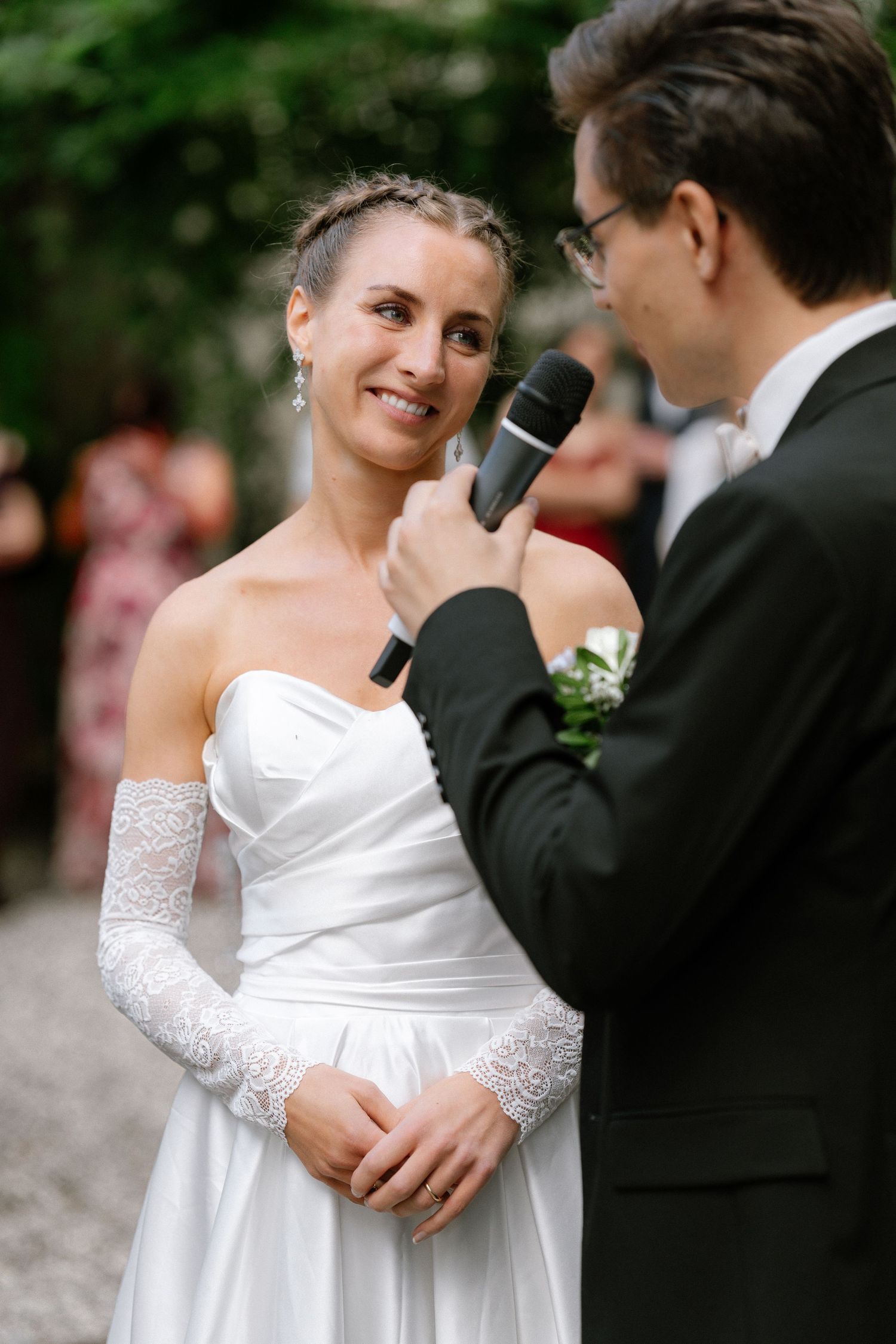 Smiling bride in a white strapless gown and lace sleeves listens as a groom in a dark suit speaks into a microphone at an outdoor wedding.