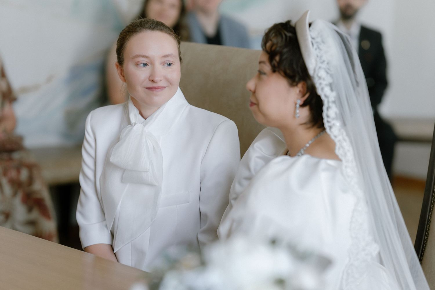 Two brides in white wedding attire share a tender moment at their ceremony, one wearing a veil.