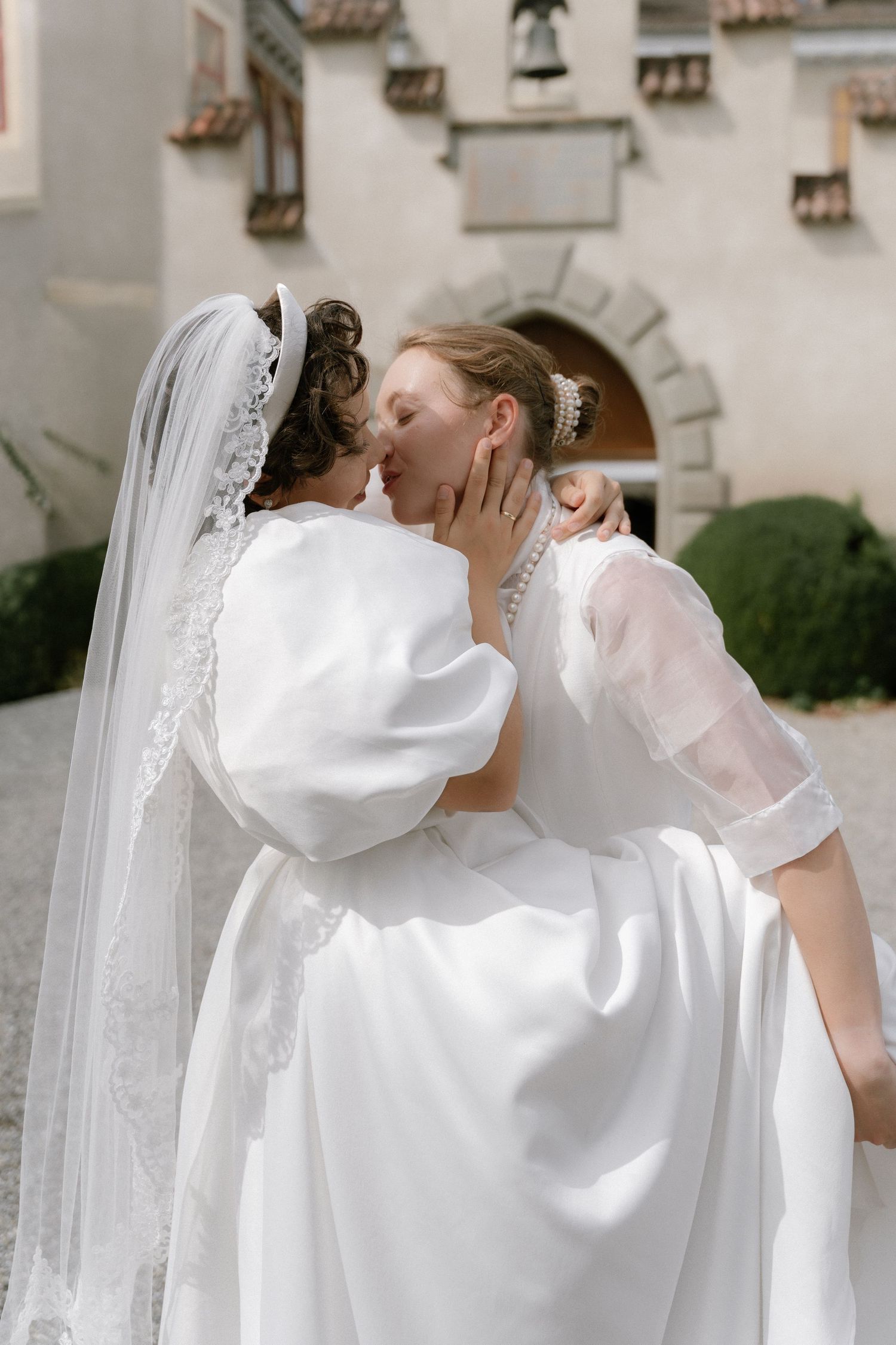 Two brides in white gowns share a romantic kiss outside a historic building on their wedding day.