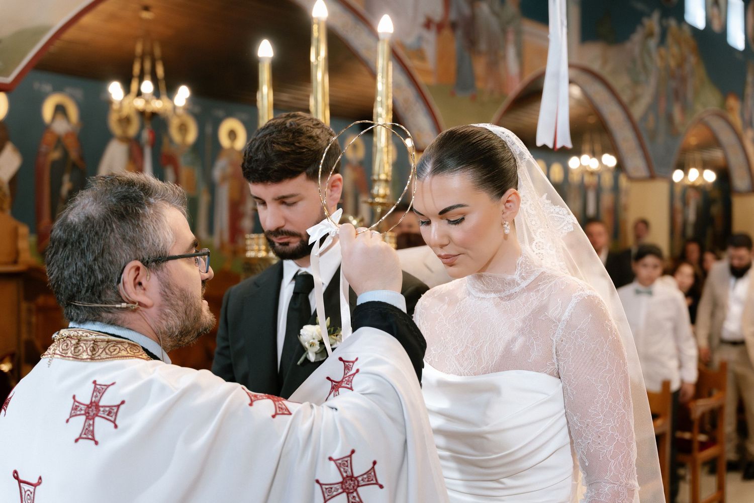 Orthodox priest crowning bride and groom during traditional Greek wedding ceremony inside ornate church.