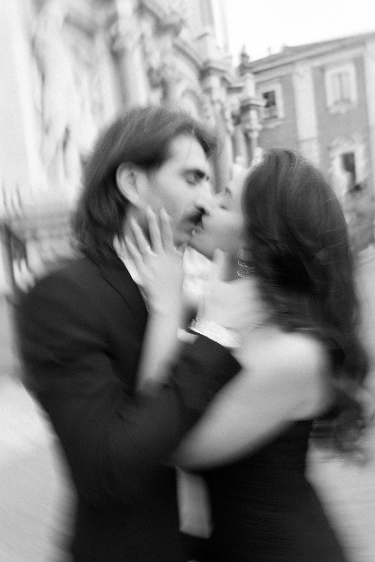 Black and white photo of a couple sharing a romantic kiss outdoors near a historic European building.