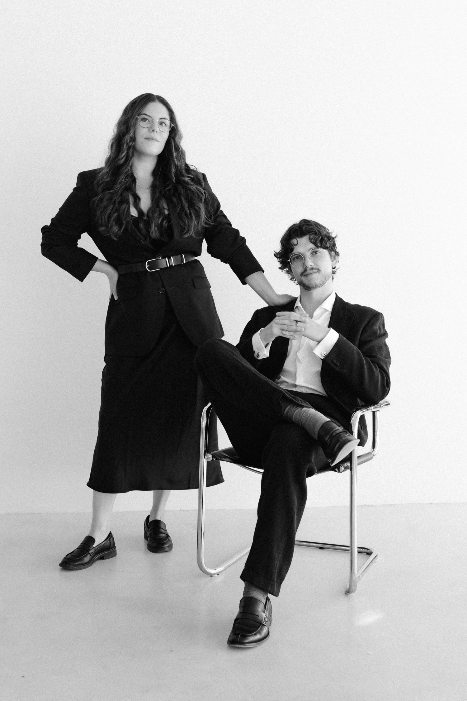 Black and white photo of a woman standing and a man seated in a chair, both wearing elegant black suits in a studio.