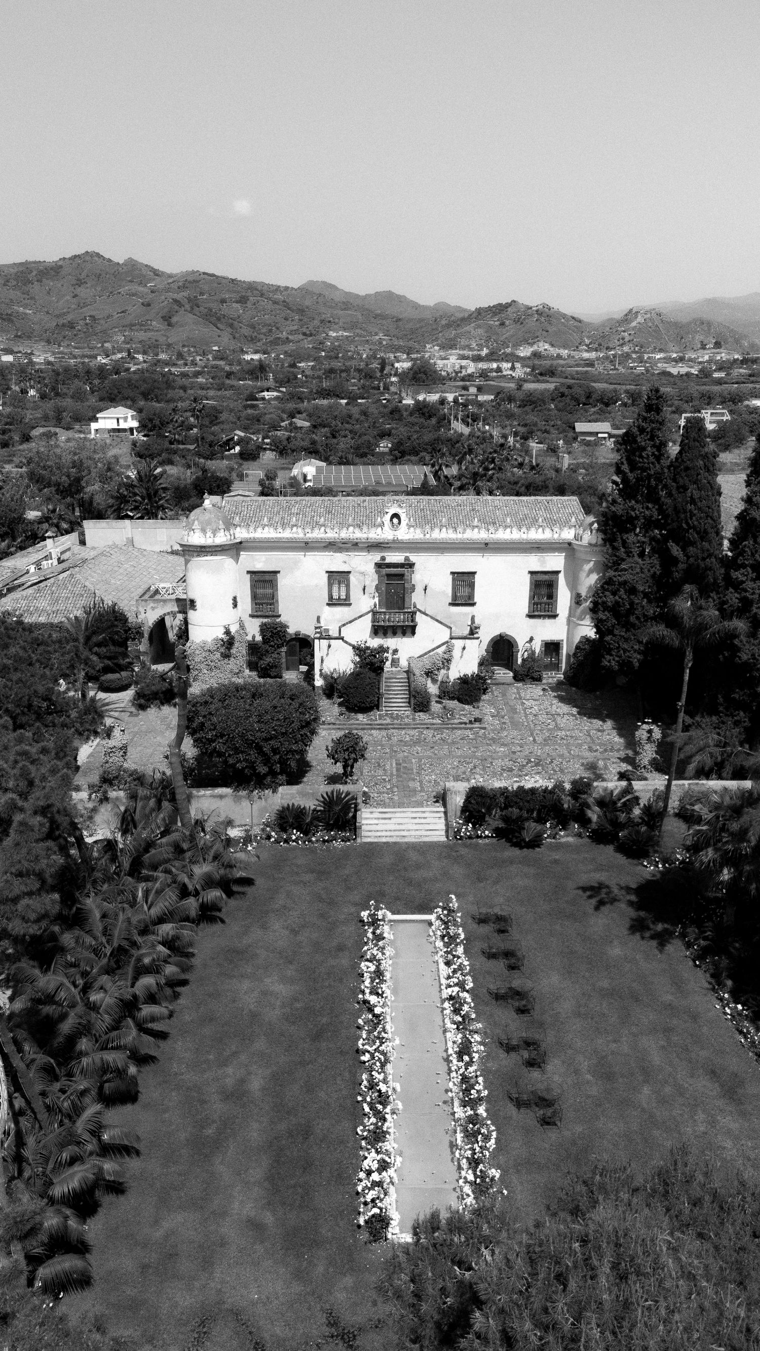 Aerial black and white view of a grand historic villa with formal gardens, fountain, and mountain backdrop.