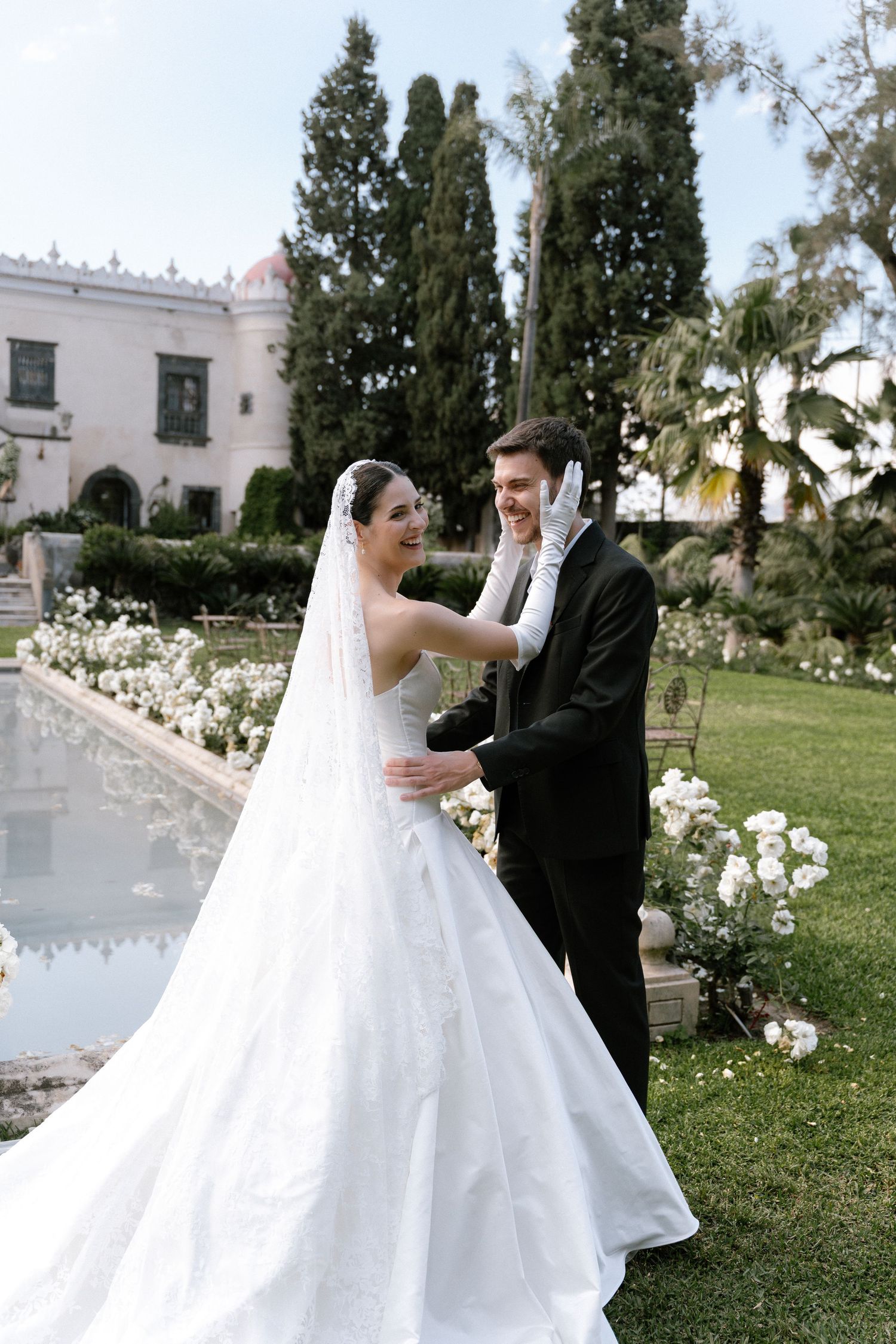 Bride in white gown and veil smiles at groom in black tuxedo in lush garden with white floral arrangements.