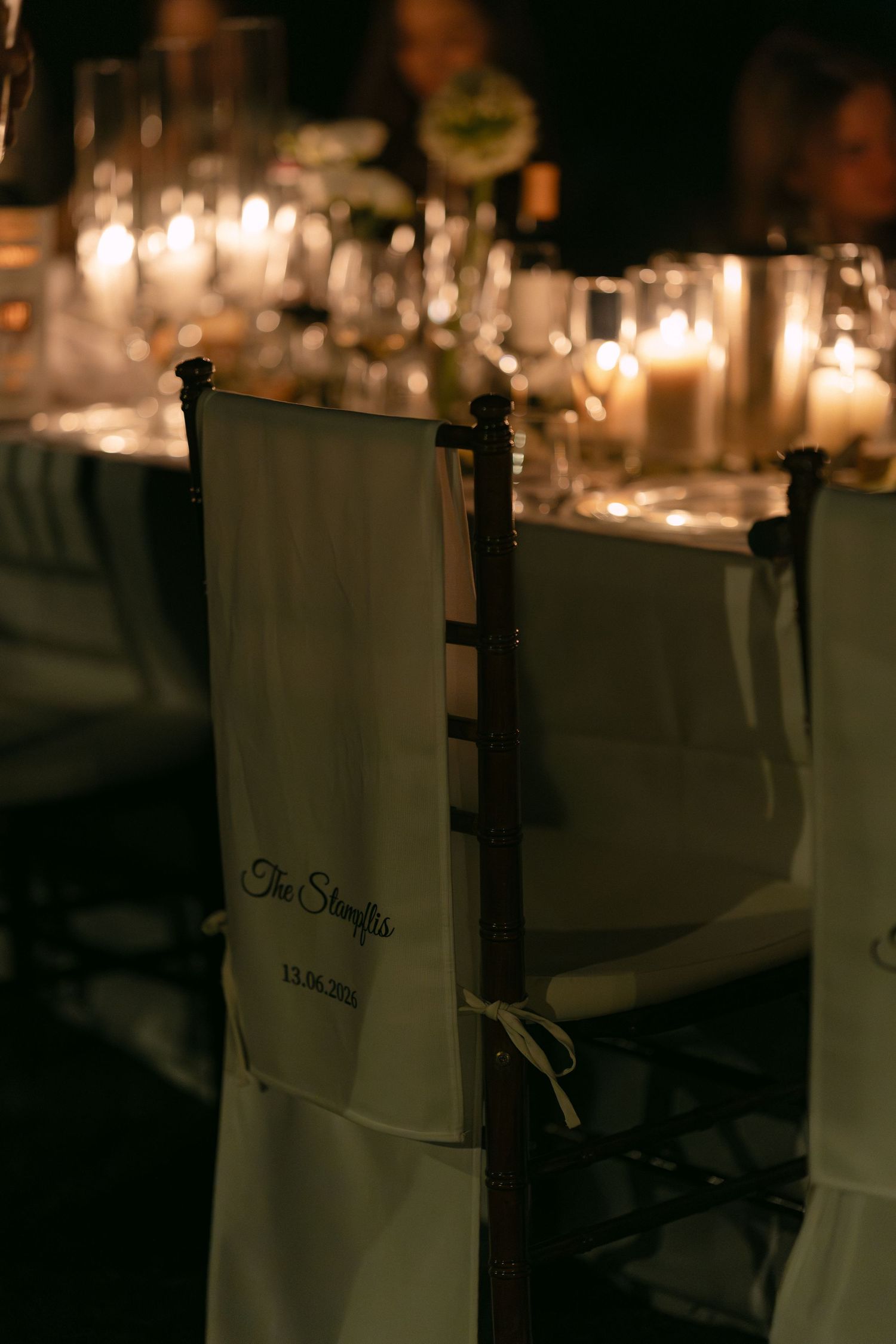 Elegant wedding reception chair with personalized white sash, candlelit table setting in warm romantic ambiance.