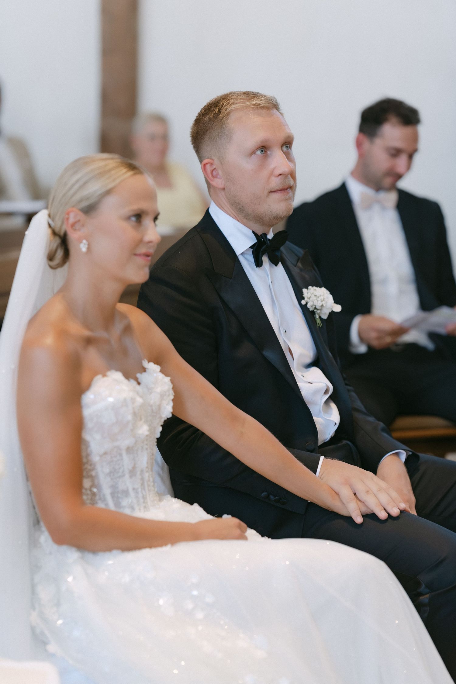 Bride in a sparkling strapless gown and groom in a black tuxedo with boutonniere sit holding hands during a wedding ceremony.