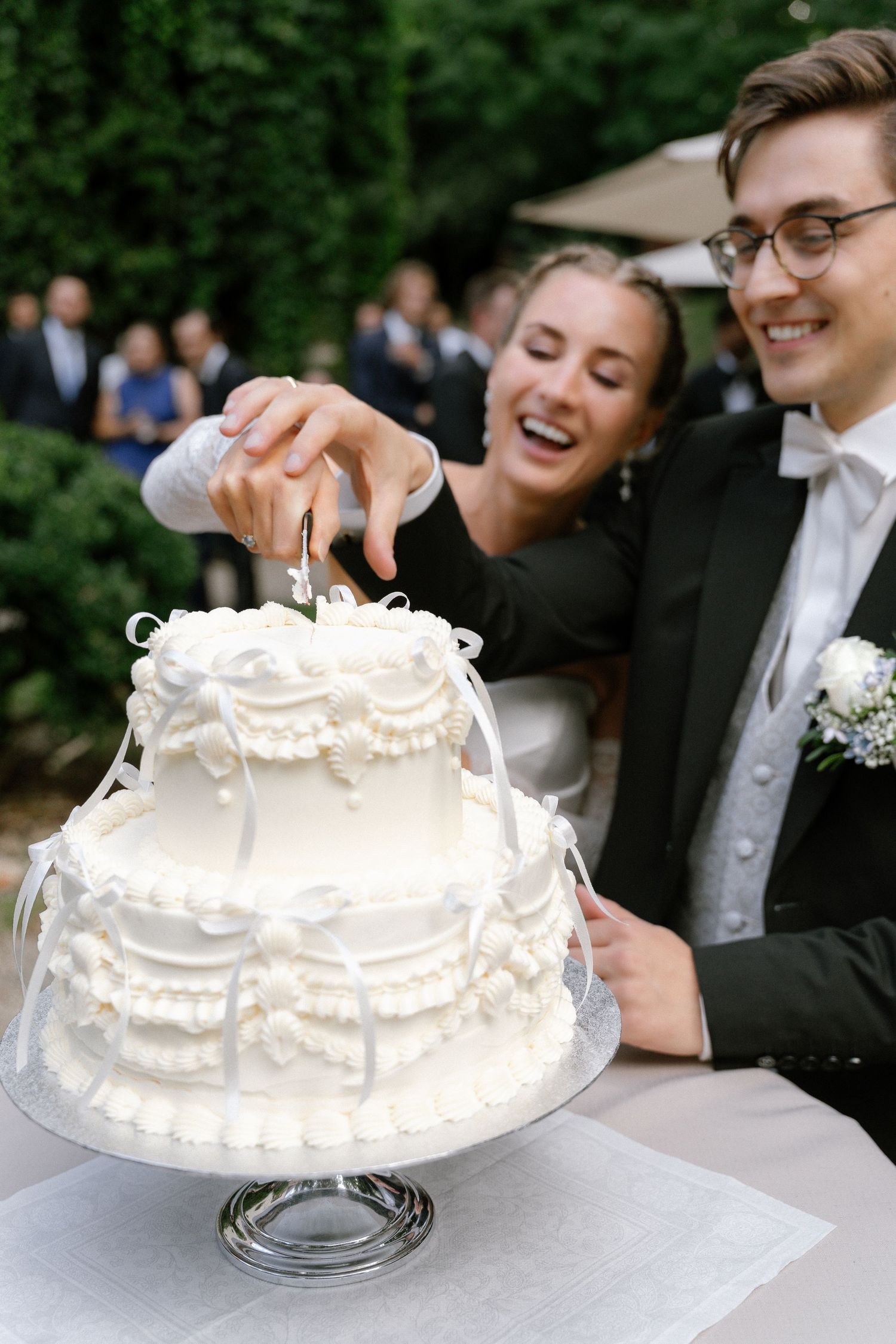 Smiling couple in formal wedding attire cutting a white vintage-style two-tier cake with ribbon details at an outdoor garden reception.