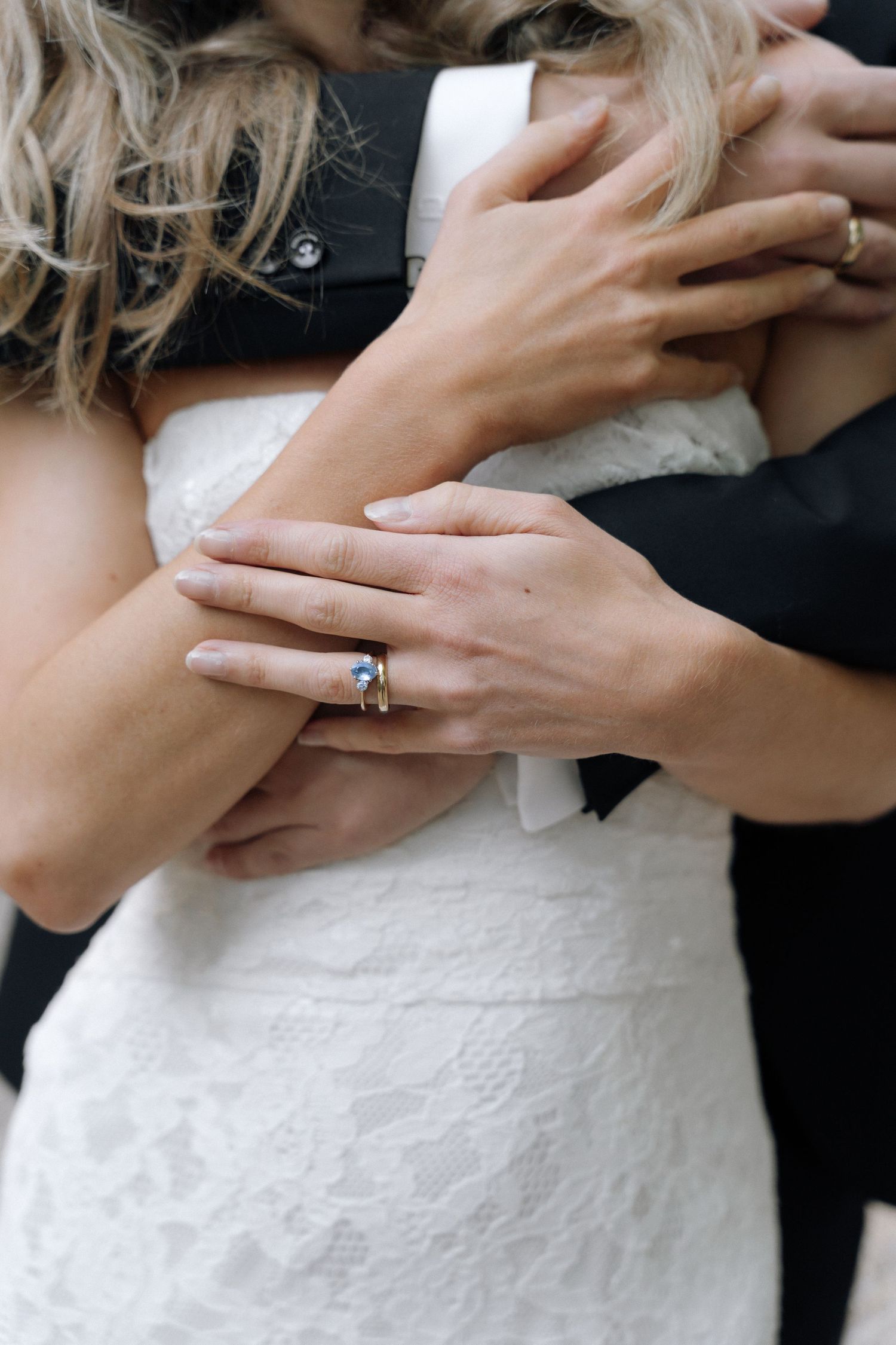 Close-up of a bride in a white lace wedding dress embraced by a groom in a dark suit, her hand with a blue gemstone ring resting on his arm.