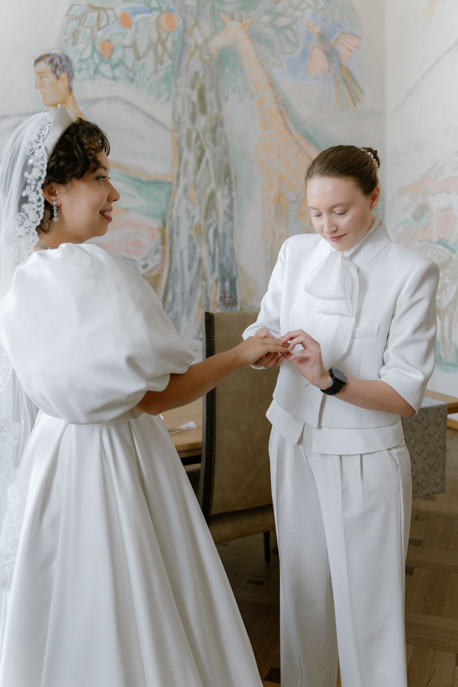 Two women in white outfits, one a bride with veil, share a tender moment in an elegant decorated room.