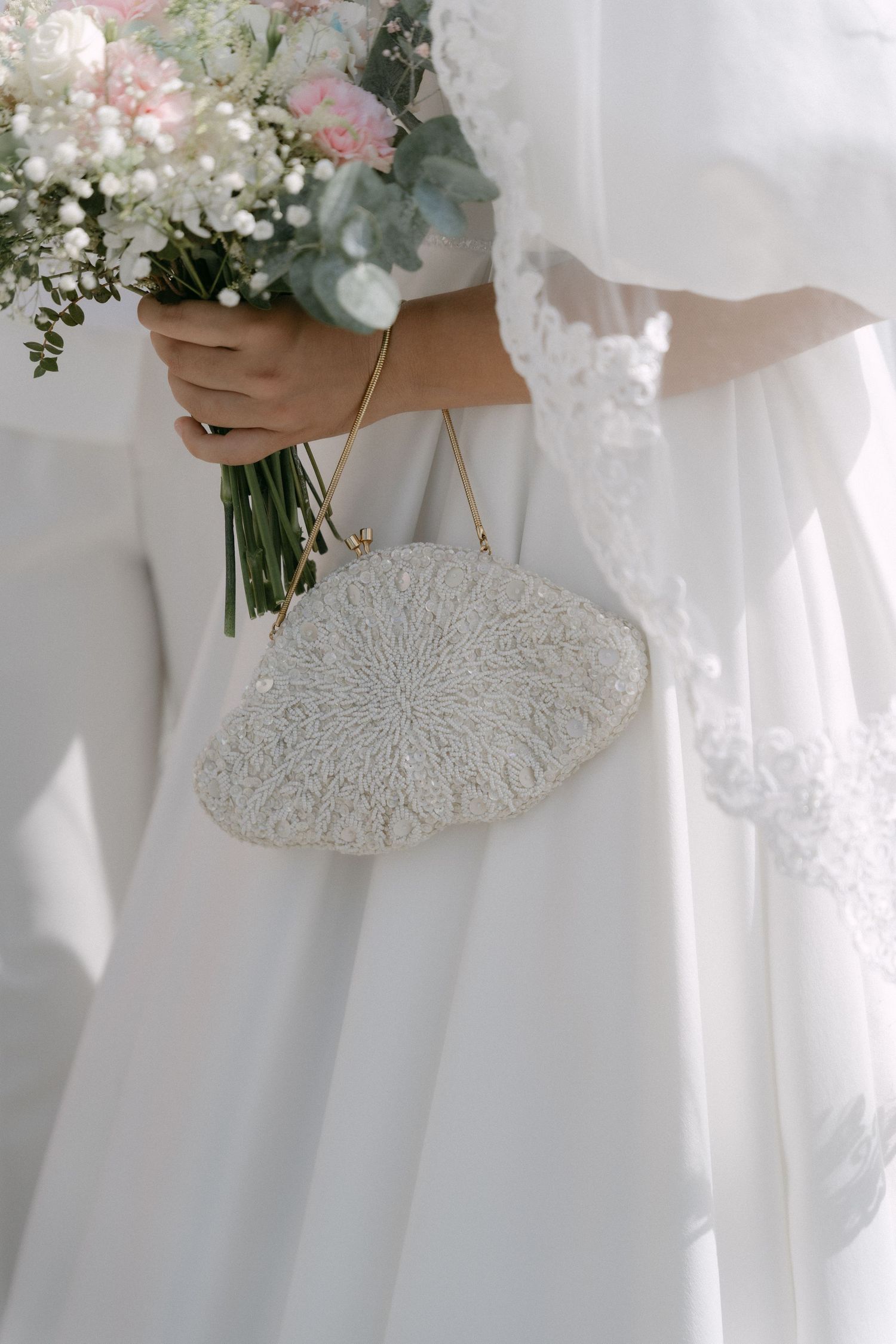 Bride holding a beaded white clutch purse and floral bouquet with pink and white flowers in a wedding dress.