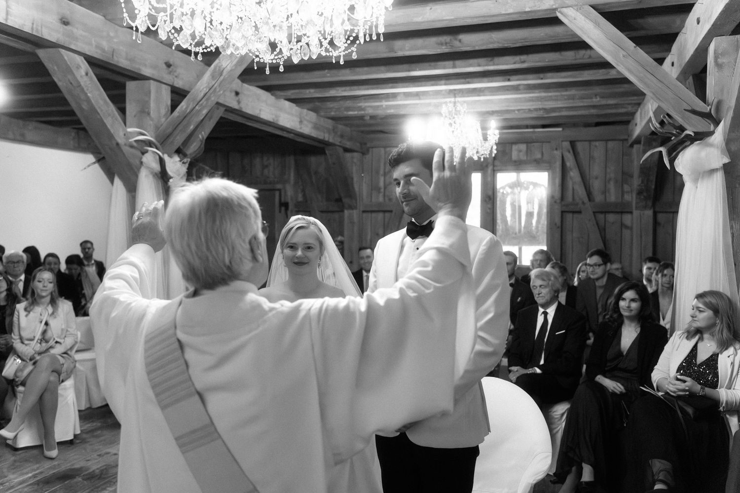 A black and white wedding ceremony photo showing people in formal attire under elegant crystal chandeliers in a rustic venue.