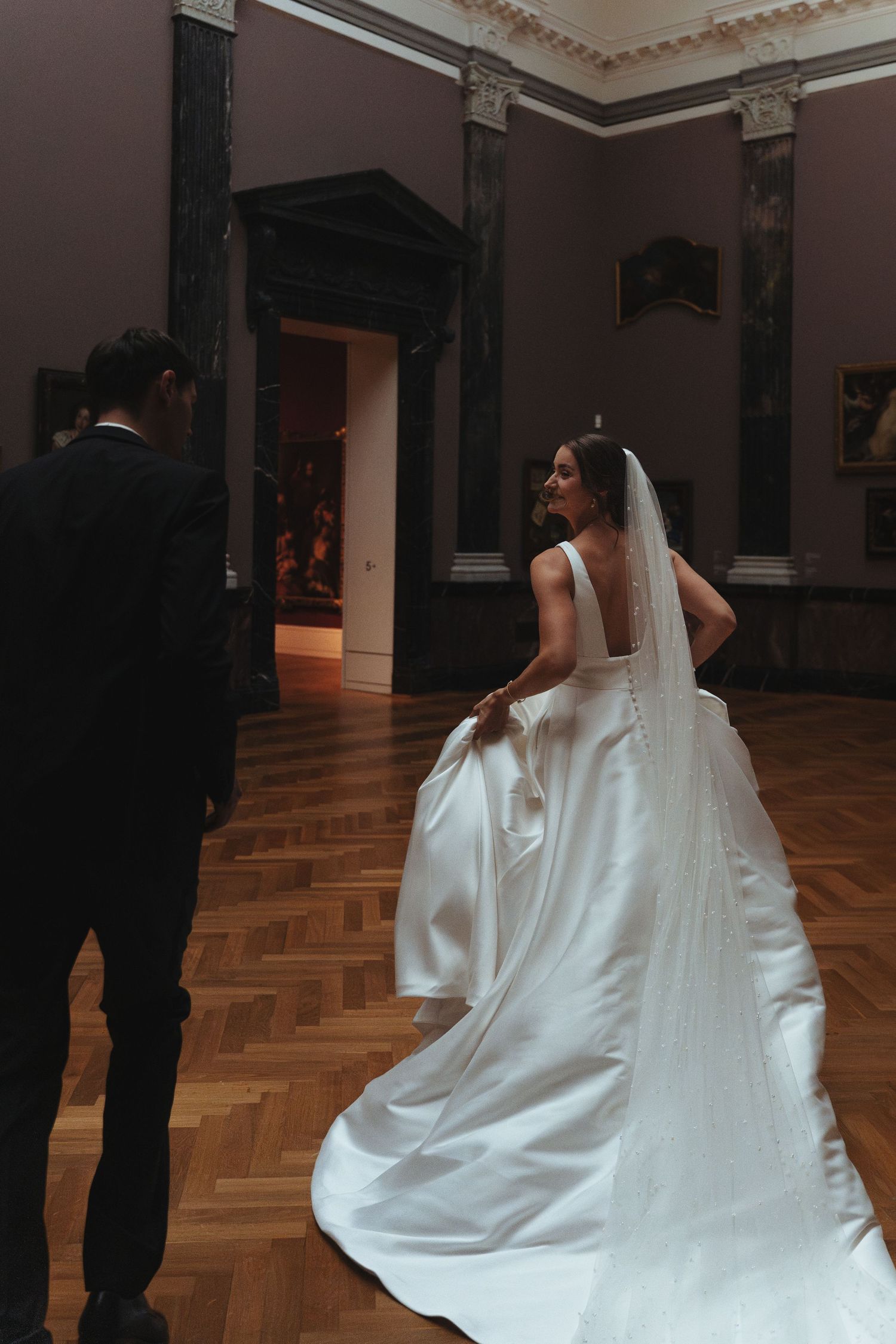 A bride in a flowing white wedding dress with a long train walks through an elegant room with parquet flooring.