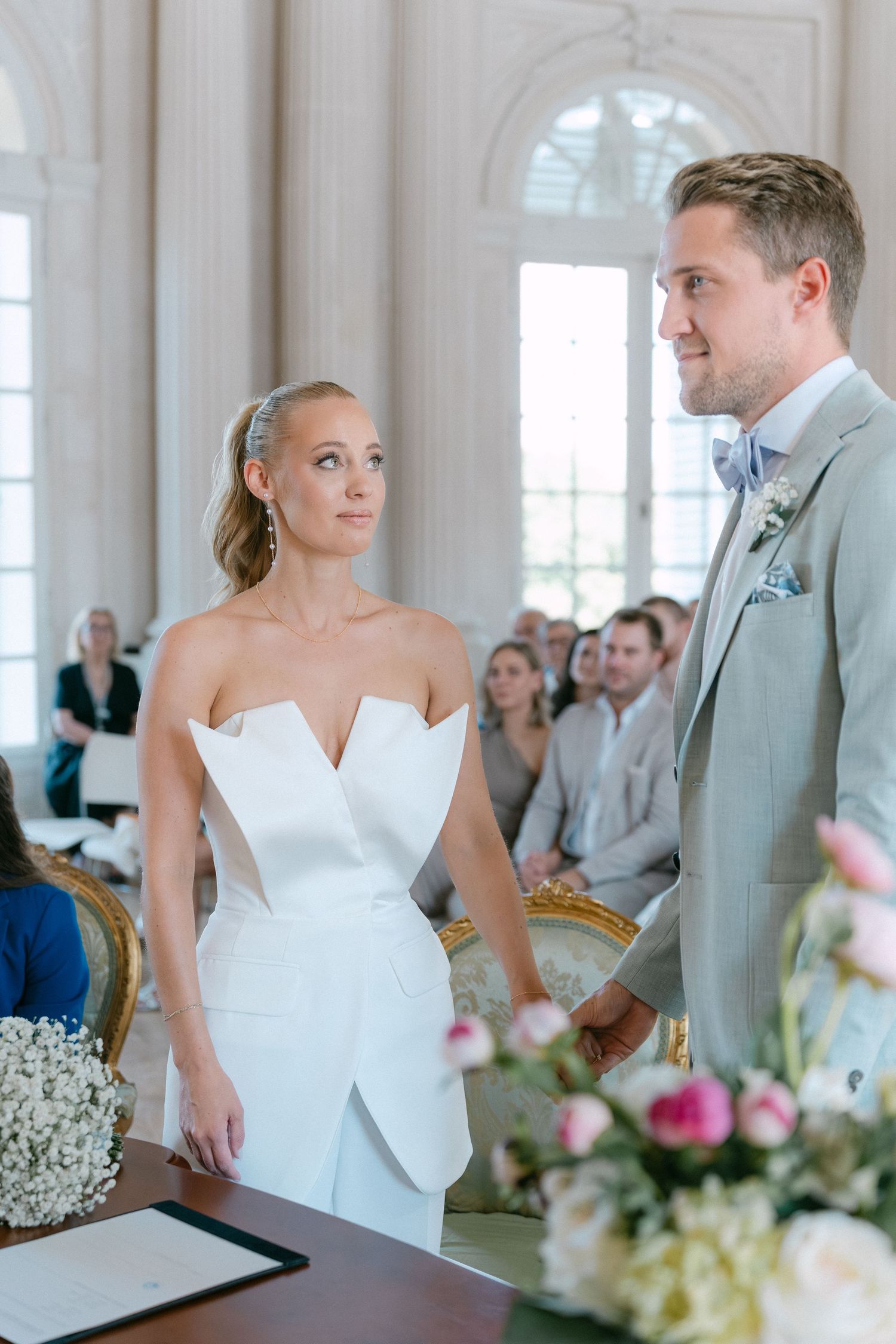 A bride in a modern white strapless dress and groom in light gray suit exchange glances during their wedding ceremony. Schloss Solitude