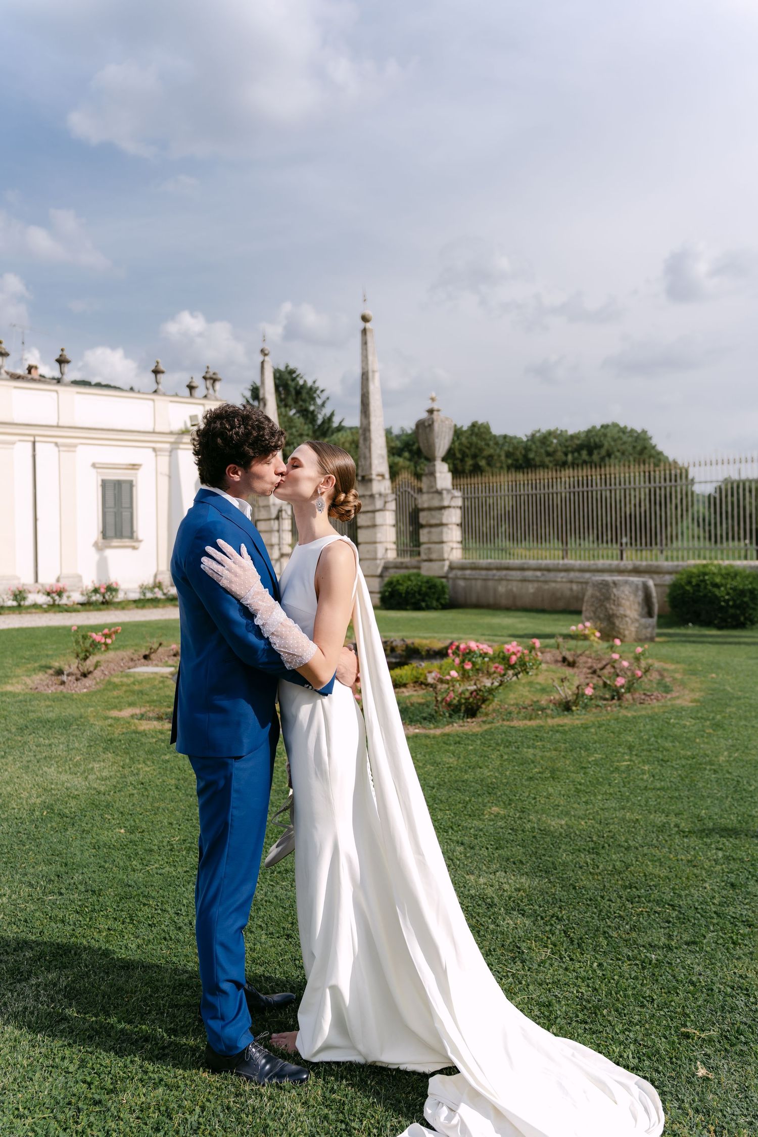 A couple shares romantic wedding moments in a garden setting with ornate stone pillars and lush green lawns. Villa Mosconi Bertani