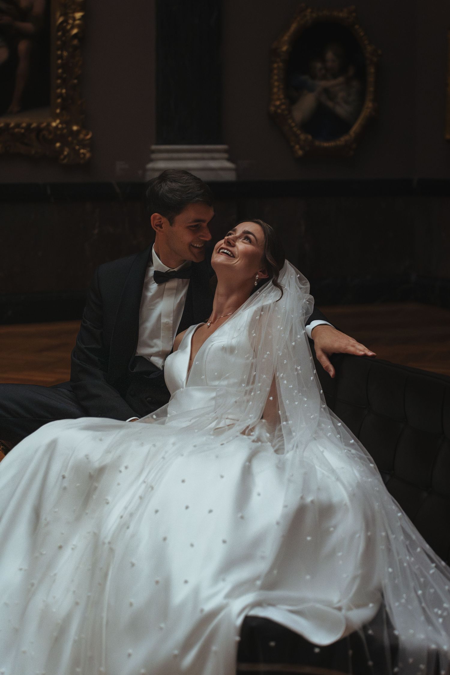 Newlywed couple shares an intimate moment on their wedding day in an elegant dark gallery with ornate decor.
