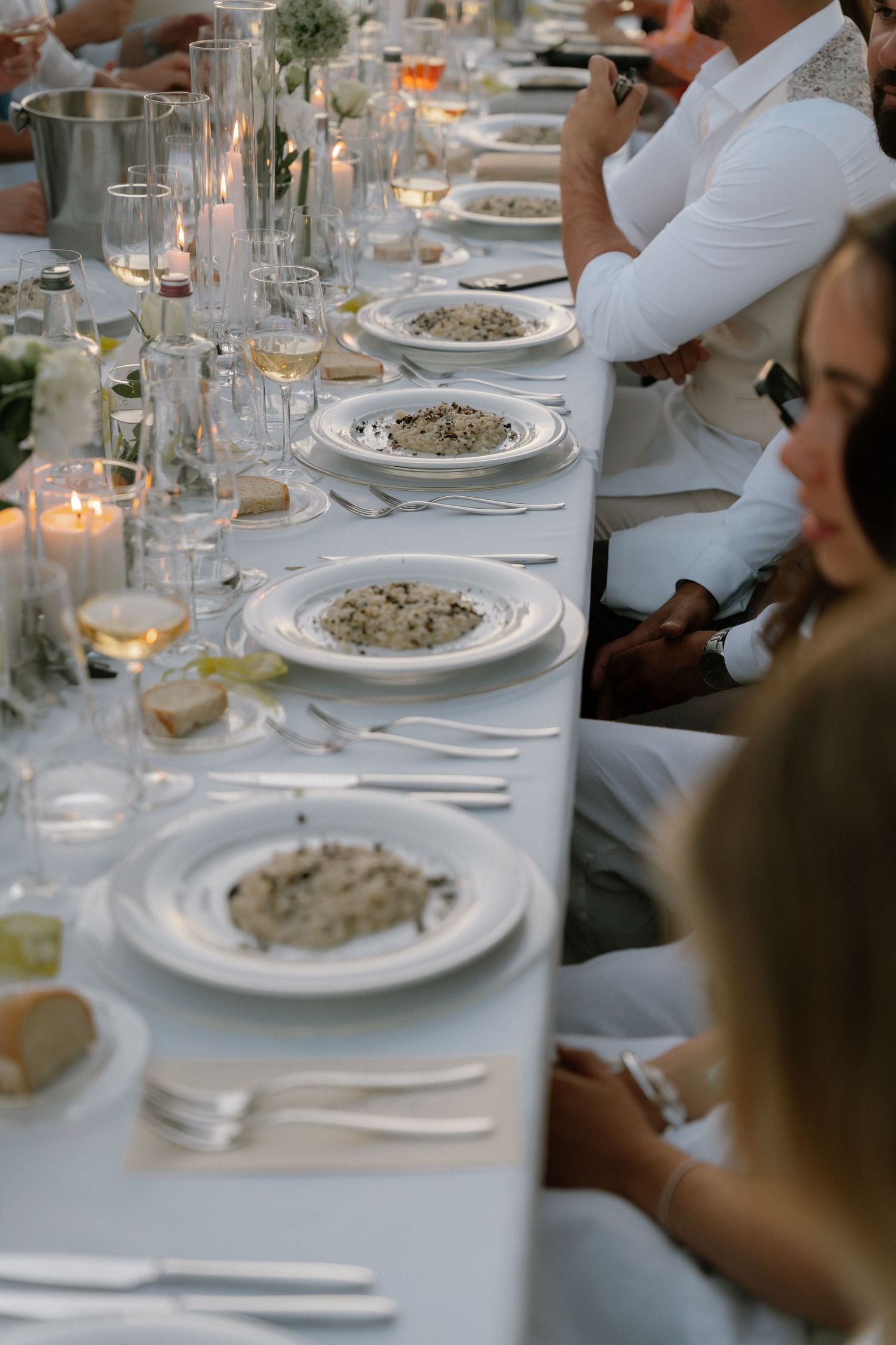 Elegant outdoor dinner party with white plates of risotto, candles, and wine glasses on a long white tablecloth.