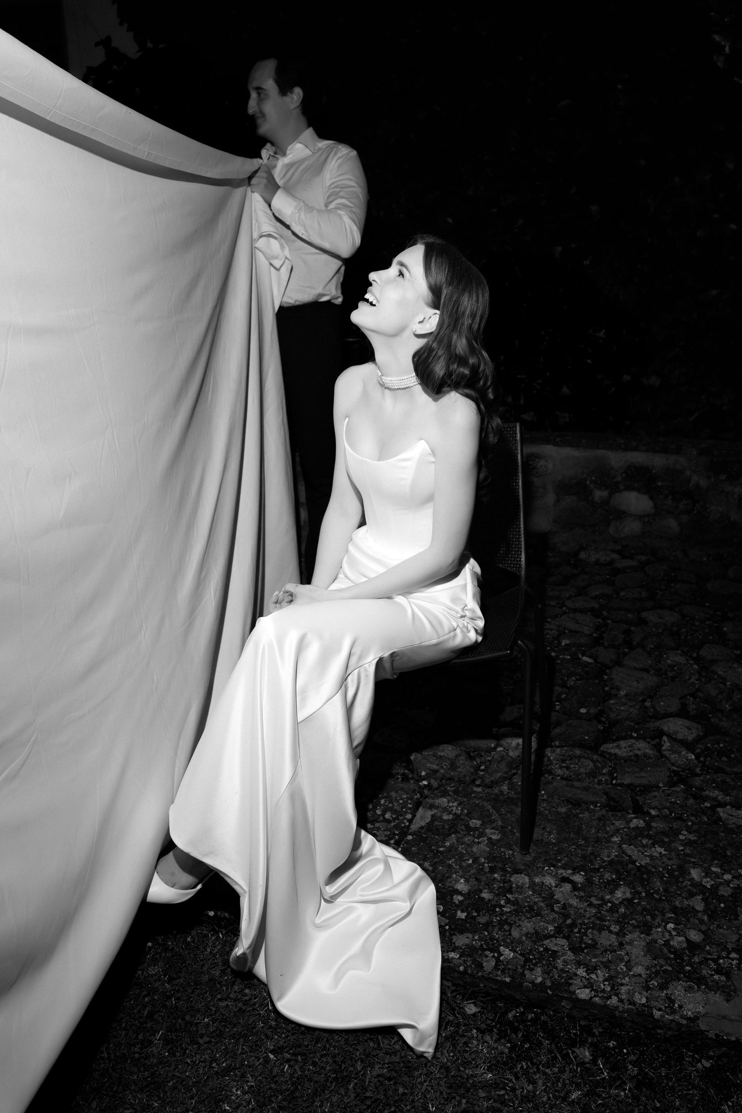 Black and white photo of a smiling bride in a satin gown seated outdoors at night, with a man holding fabric behind her.