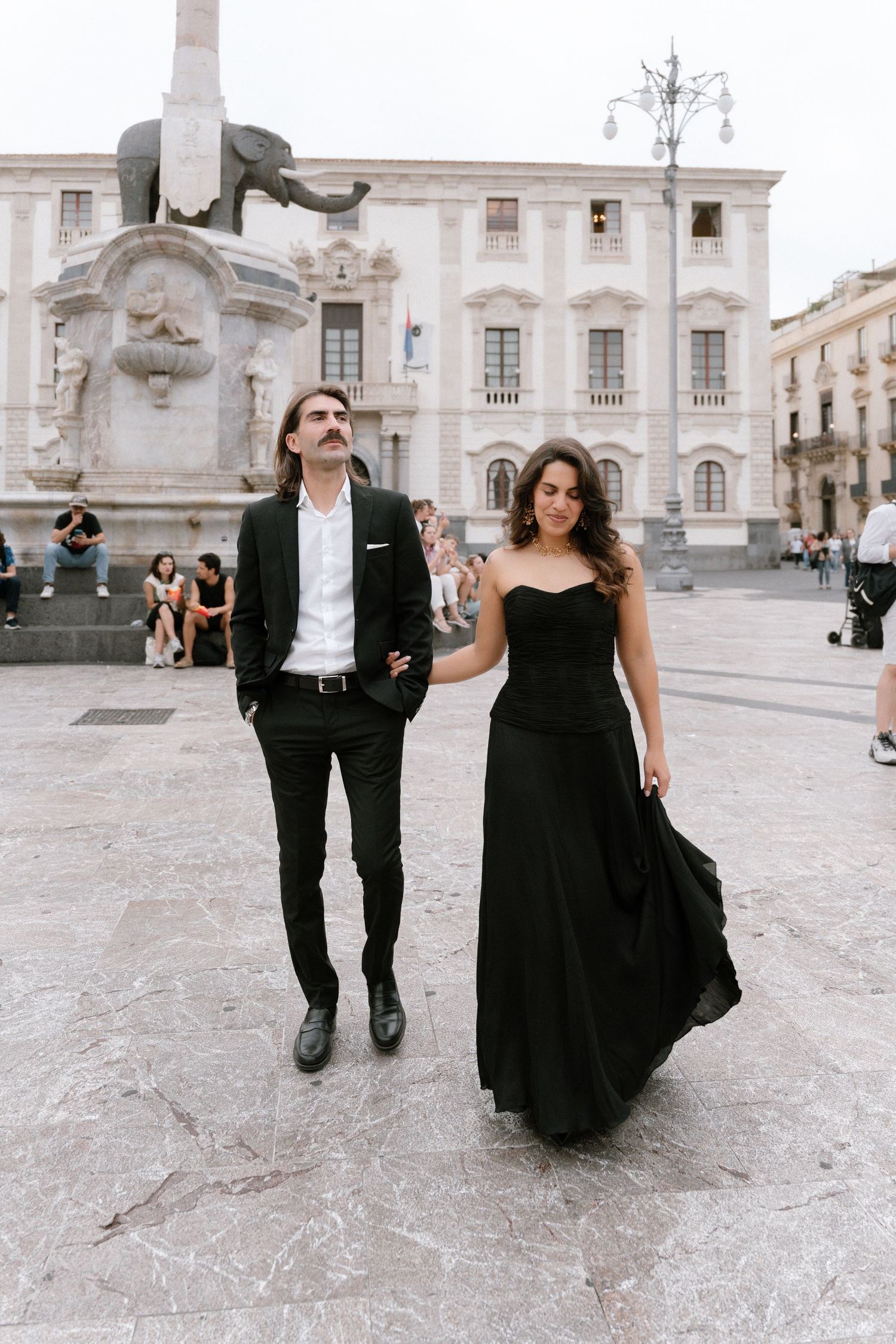Elegant couple in matching black outfits walking hand in hand in a European piazza with a fountain statue backdrop.