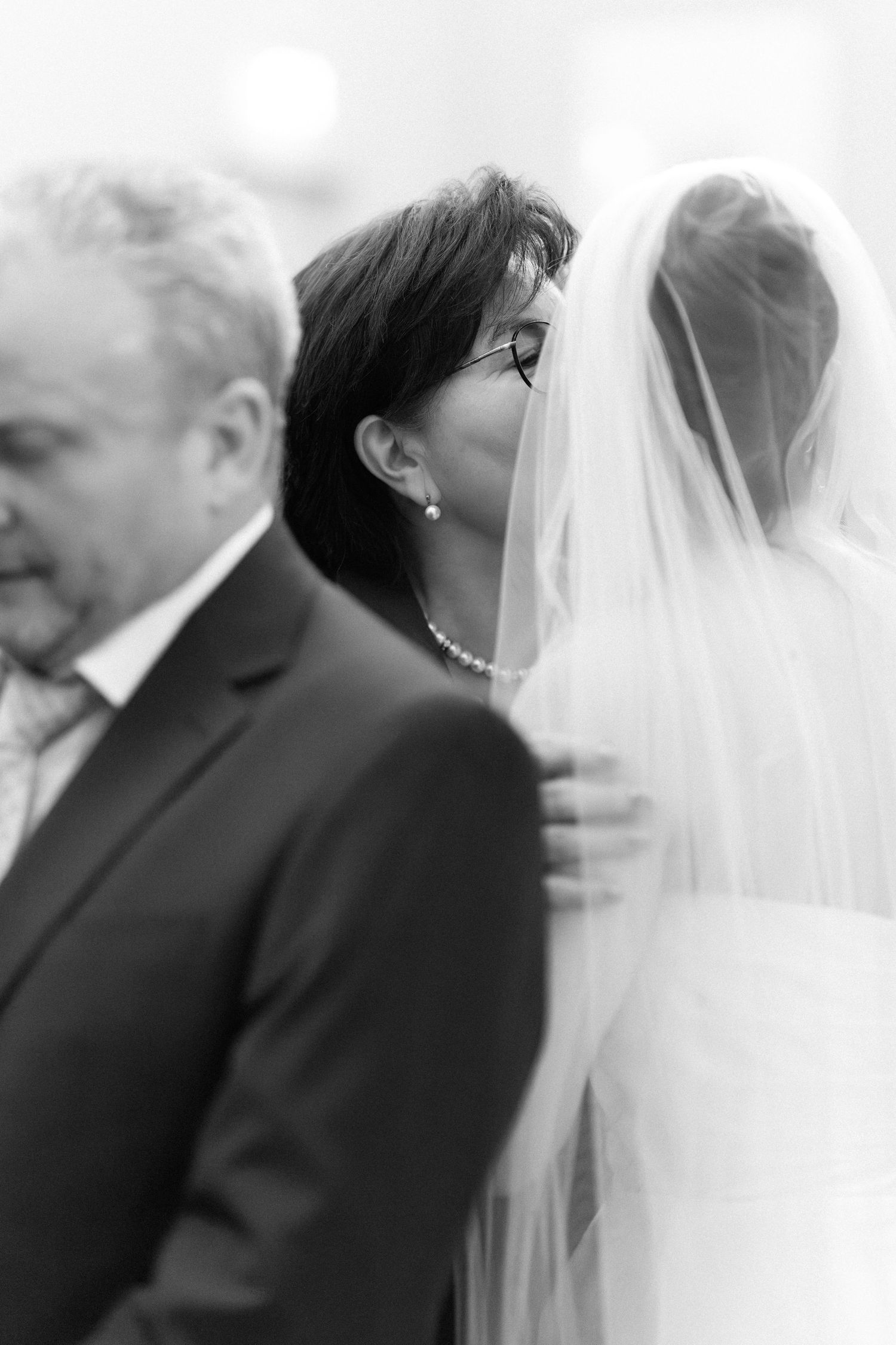 Black and white wedding photo of bride with veil, emotional mother and father standing beside her during ceremony.