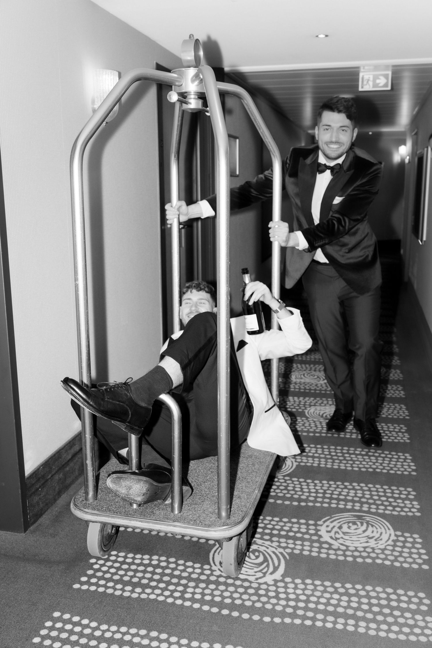 Playful black and white photo of people in formal wear posing humorously with a hotel luggage cart in a hallway.