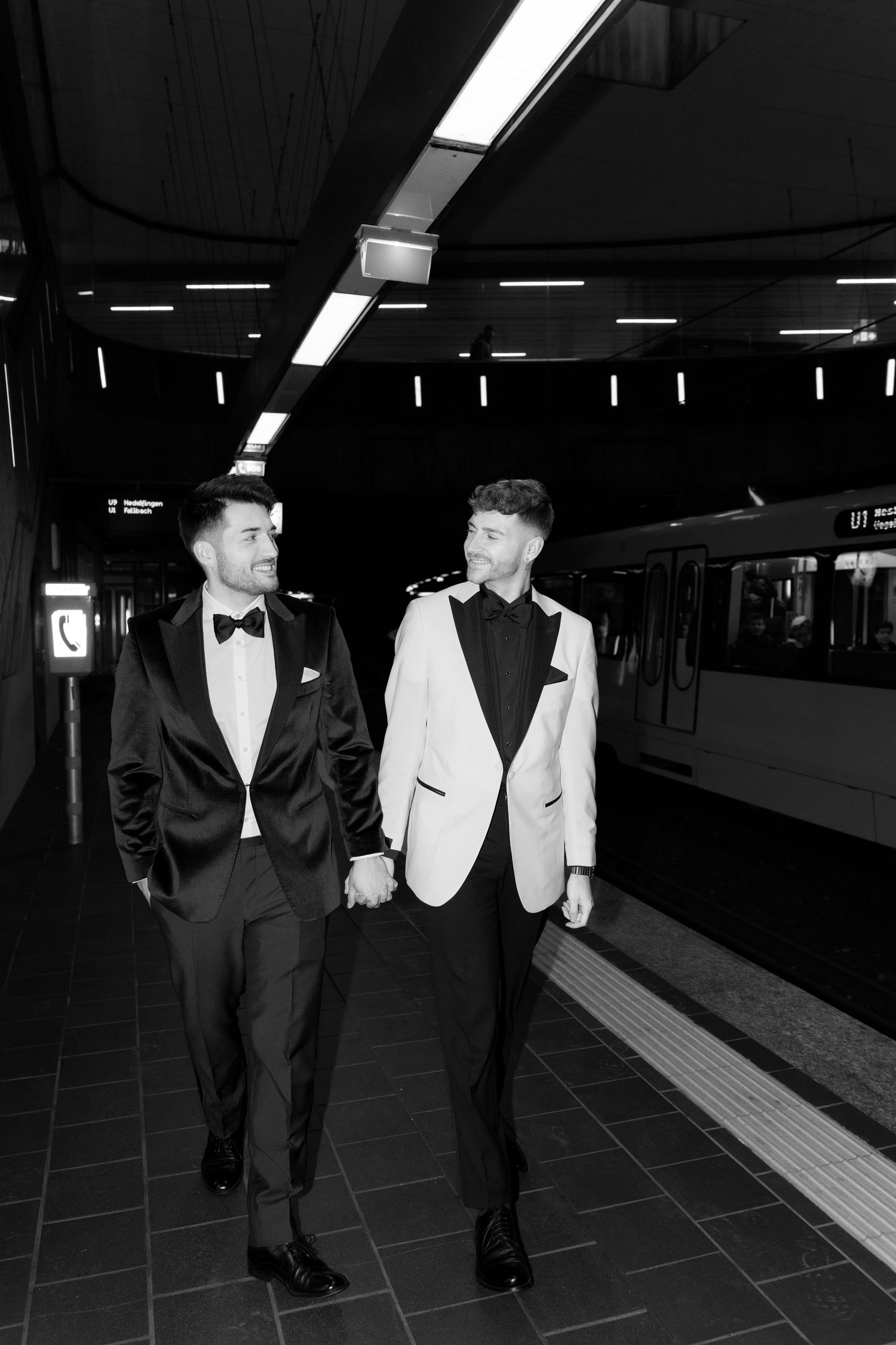 Two people in formal attire walk together through a dimly lit subway station, one wearing black and one in white.