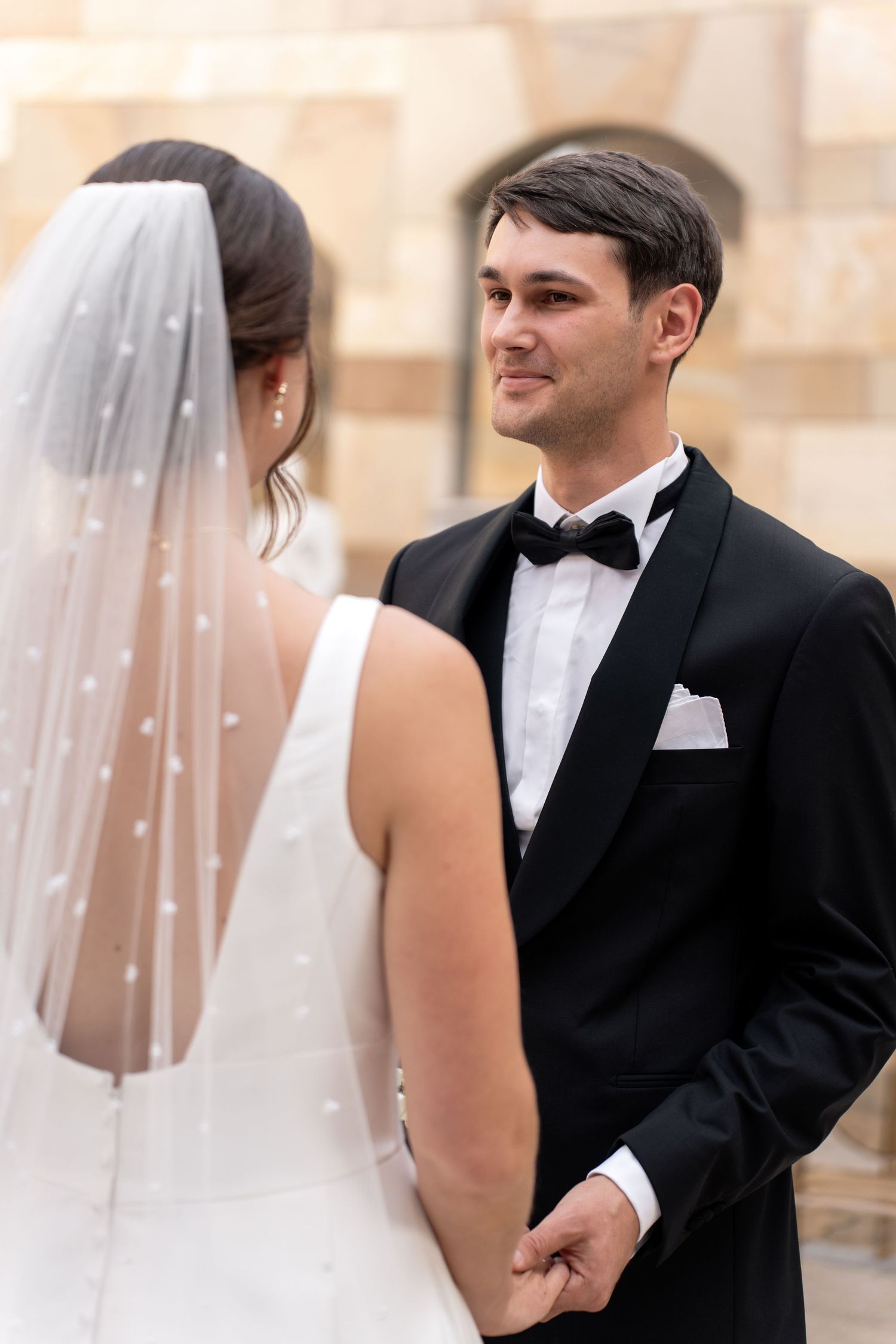 A wedding couple shares a tender moment outdoors in formal black and white attire with a flowing veil.
