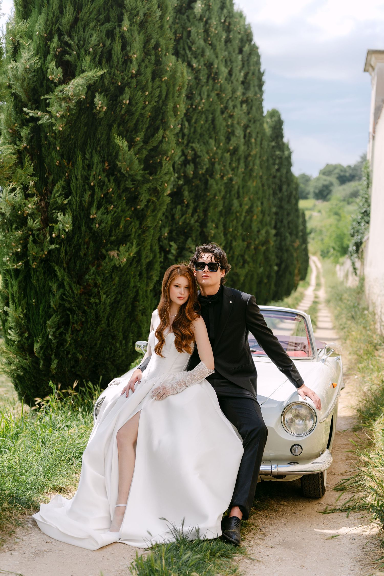 A romantic wedding couple poses with a vintage white car along a cypress tree-lined path in the countryside.