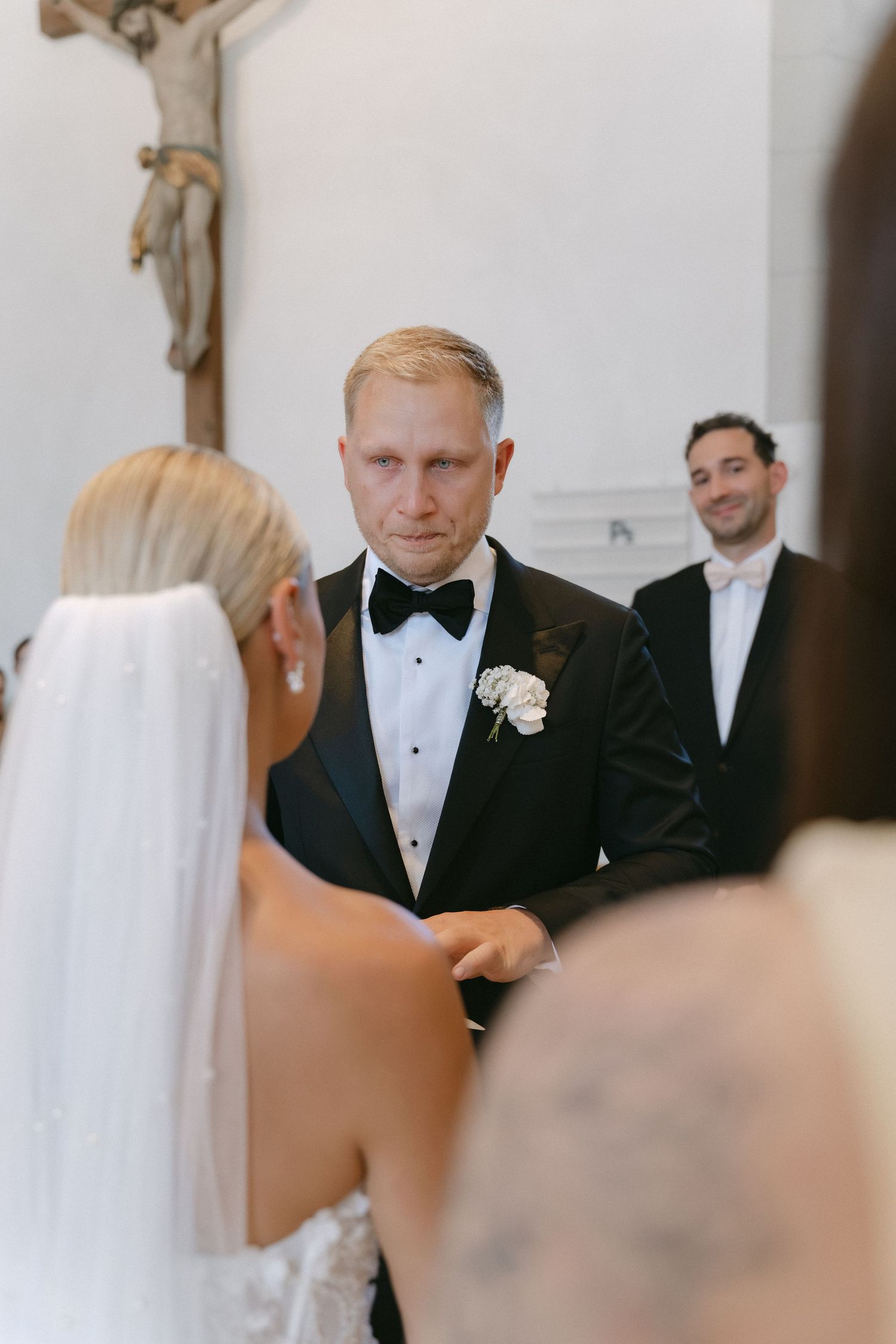 Emotional groom in a black tuxedo and bow tie faces his veiled bride during a church wedding ceremony as a smiling guest looks on.