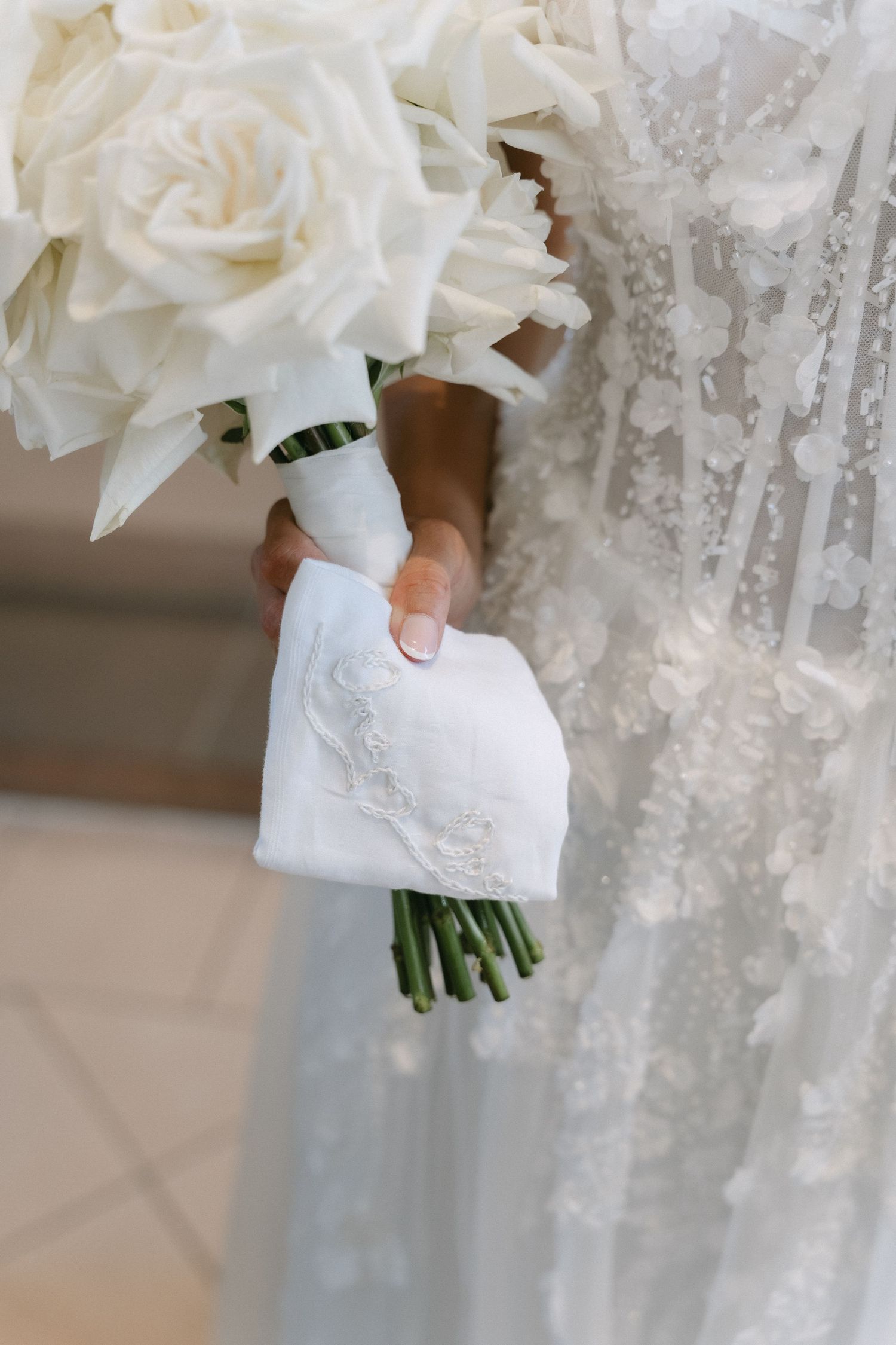 Bride in a lace floral gown holds a white rose bouquet wrapped in a white ribbon with an embroidered handkerchief tied to the stems.