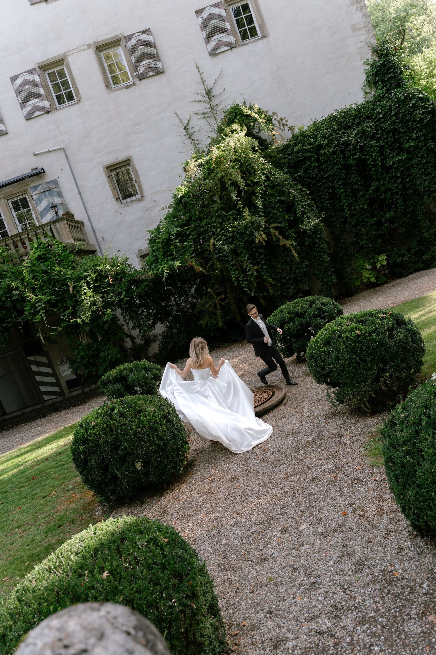 Bride in a flowing white gown walks with a groom in a dark suit through a gravel garden with trimmed boxwood shrubs beside a historic manor.