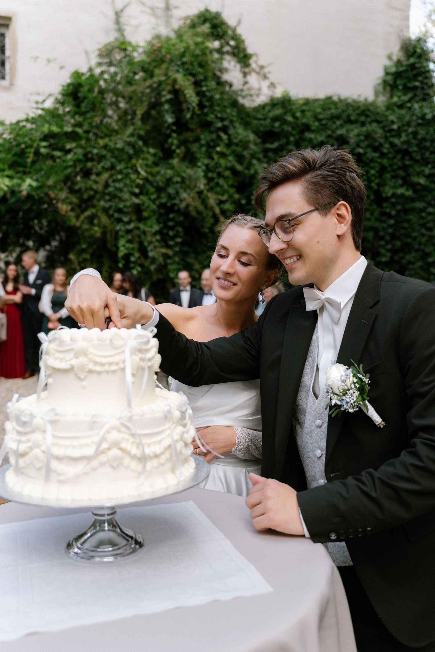 Smiling bride and groom in a black suit and bow tie cut their white tiered wedding cake together at an outdoor garden reception.