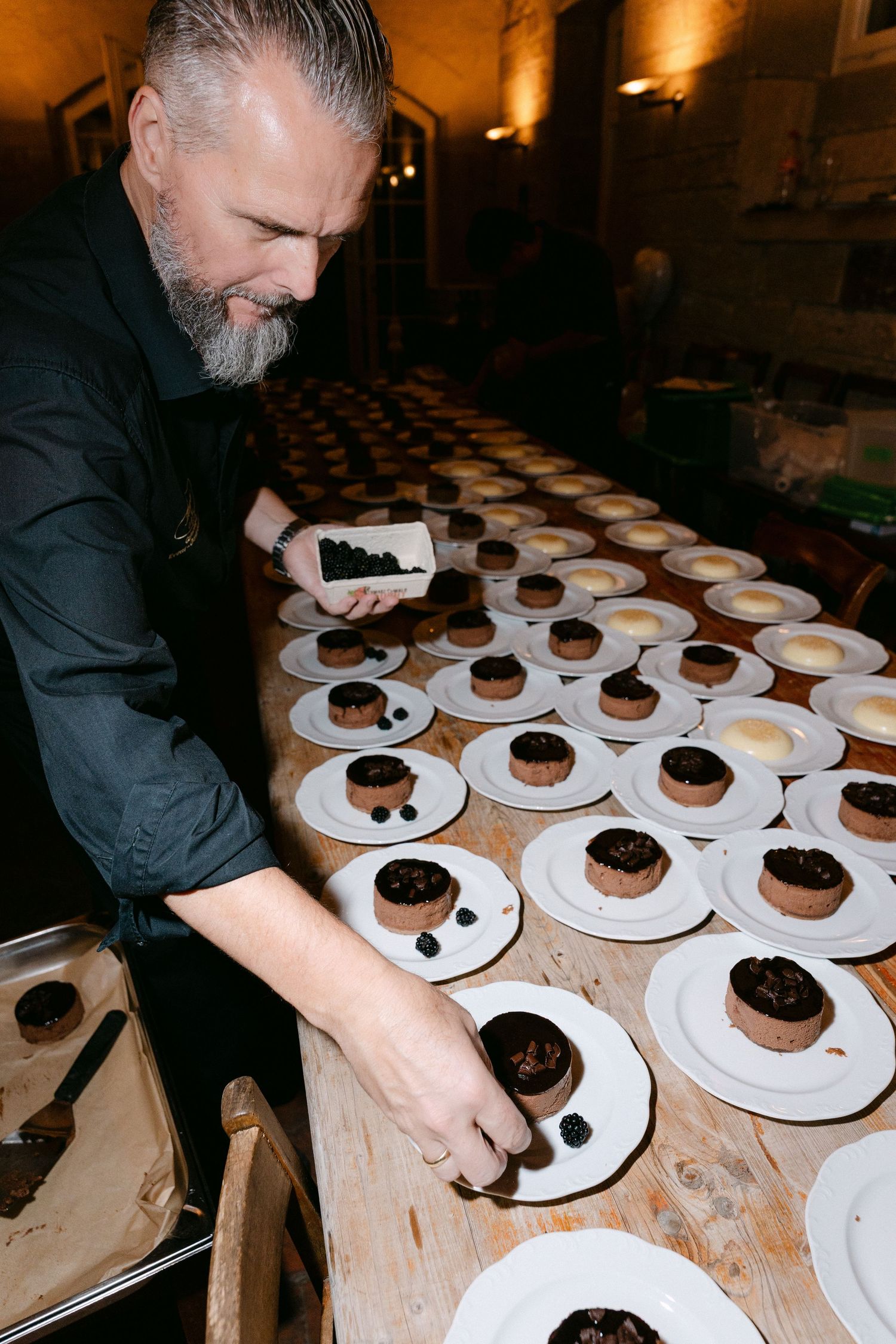 Bearded chef in a dark shirt plates dessert onto a long wooden table lined with white plates of chocolate cakes and garnishes.