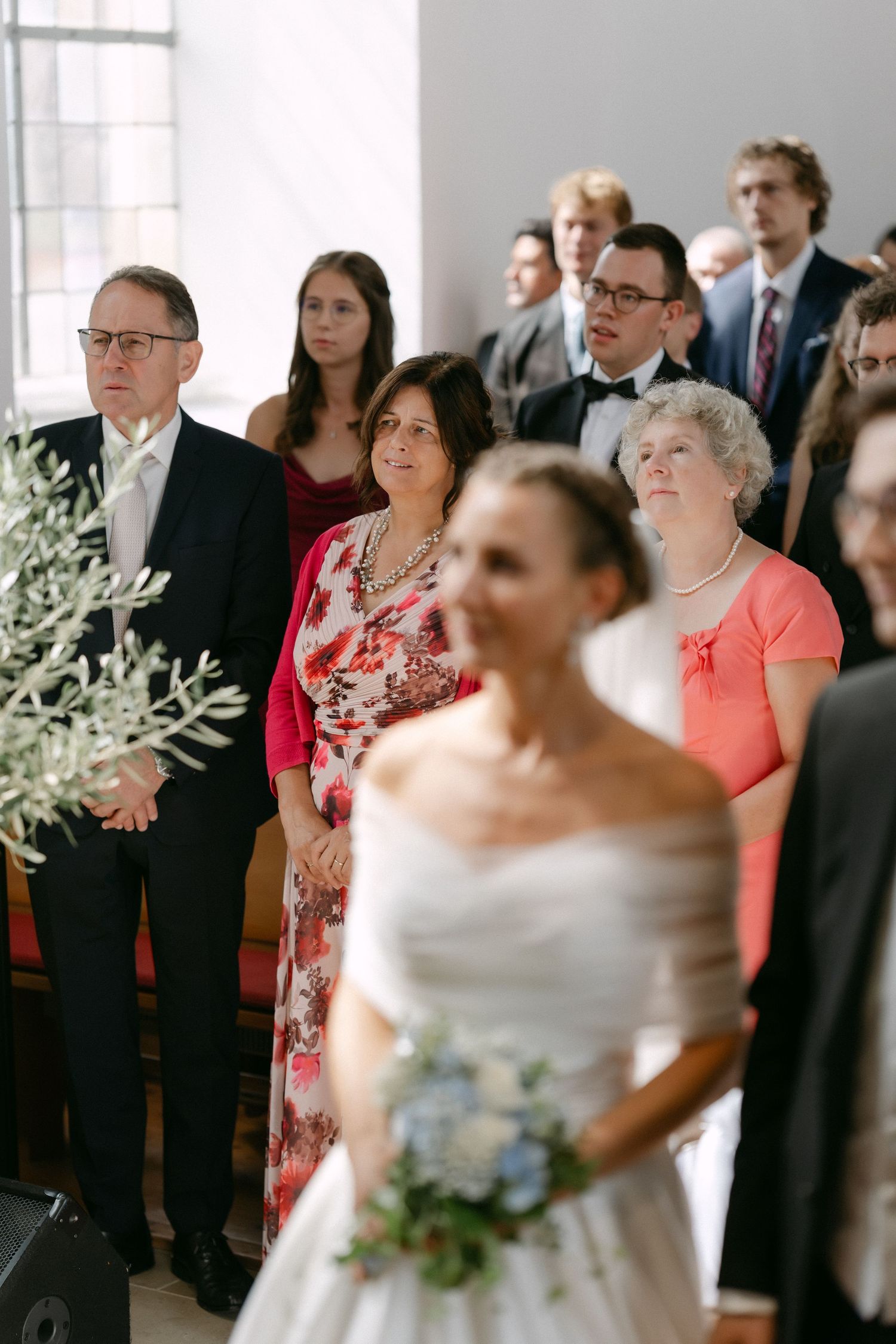 Bride in an off-shoulder white gown holding a blue floral bouquet as smiling wedding guests in formal attire watch during the ceremony.