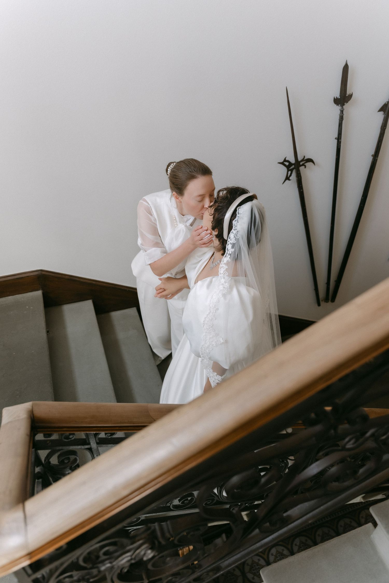 Two brides share a tender kiss on an elegant staircase, one wearing a lace wedding gown with veil.