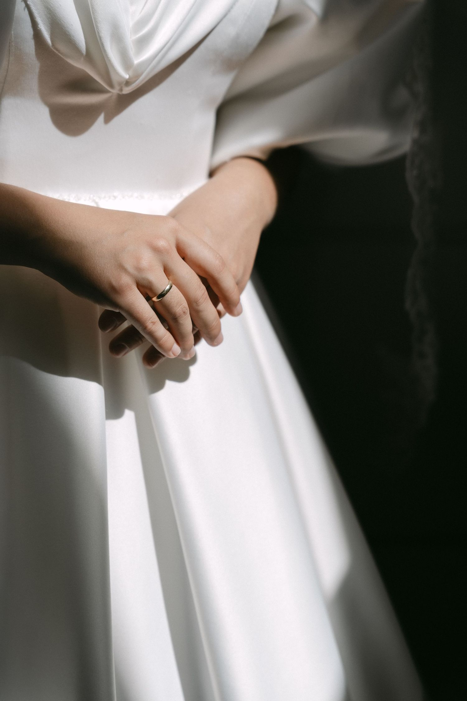 Bride in white satin wedding dress with clasped hands showing a gold wedding ring, soft natural light.