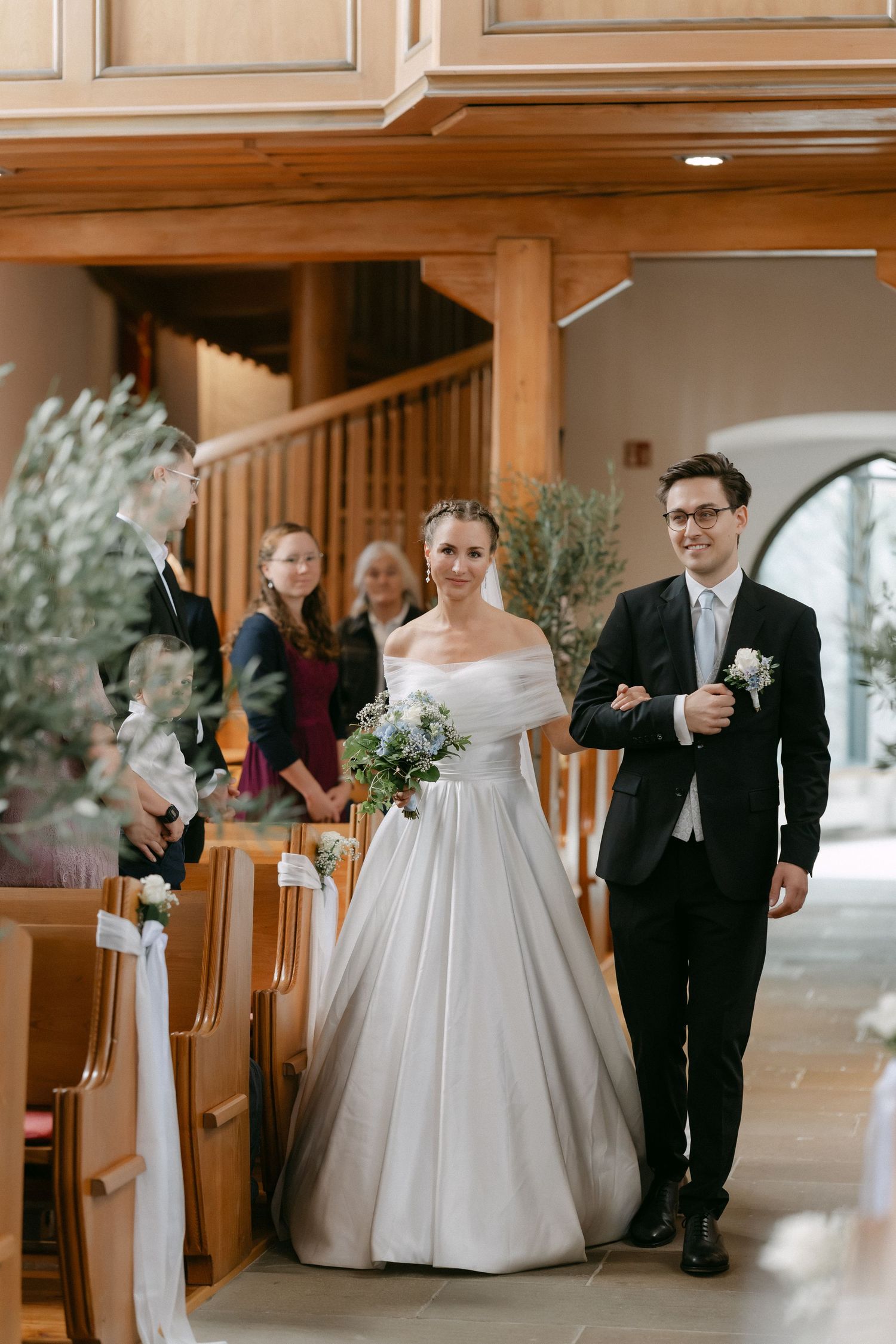 Smiling bride in an off-the-shoulder gown and groom in a dark suit walk arm in arm down the church aisle as wedding guests look on.