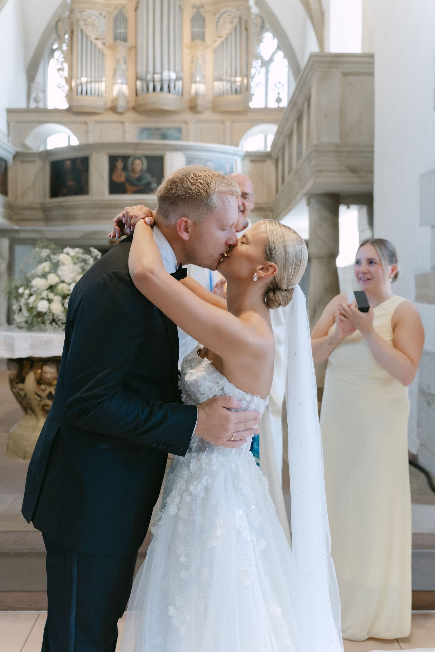 Bride in a lace wedding gown and groom in a dark suit share a kiss in a church as a bridesmaid in a yellow dress looks on.
