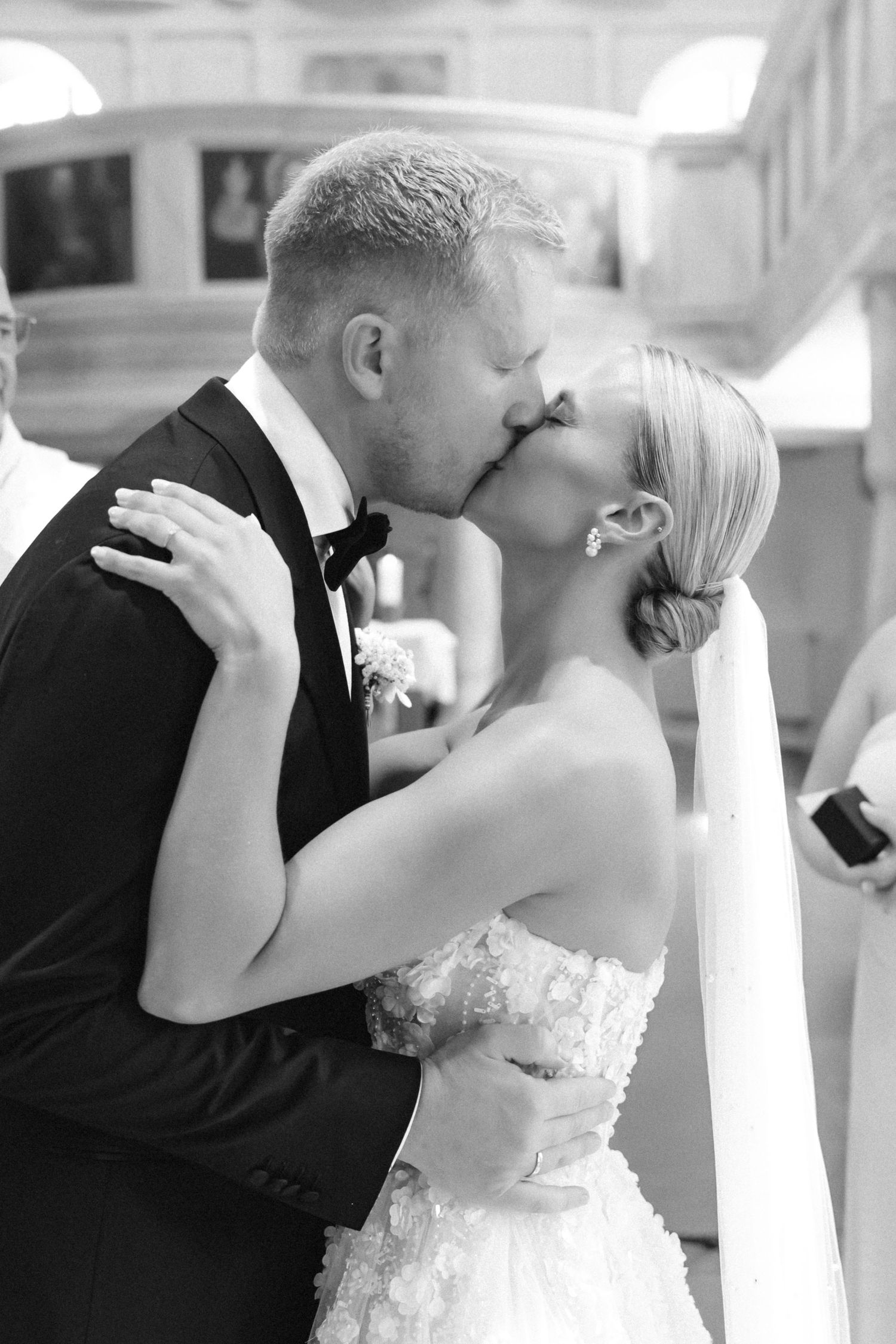Black and white photo of a bride in a strapless lace gown and a groom in a tuxedo sharing a kiss during their church wedding ceremony.