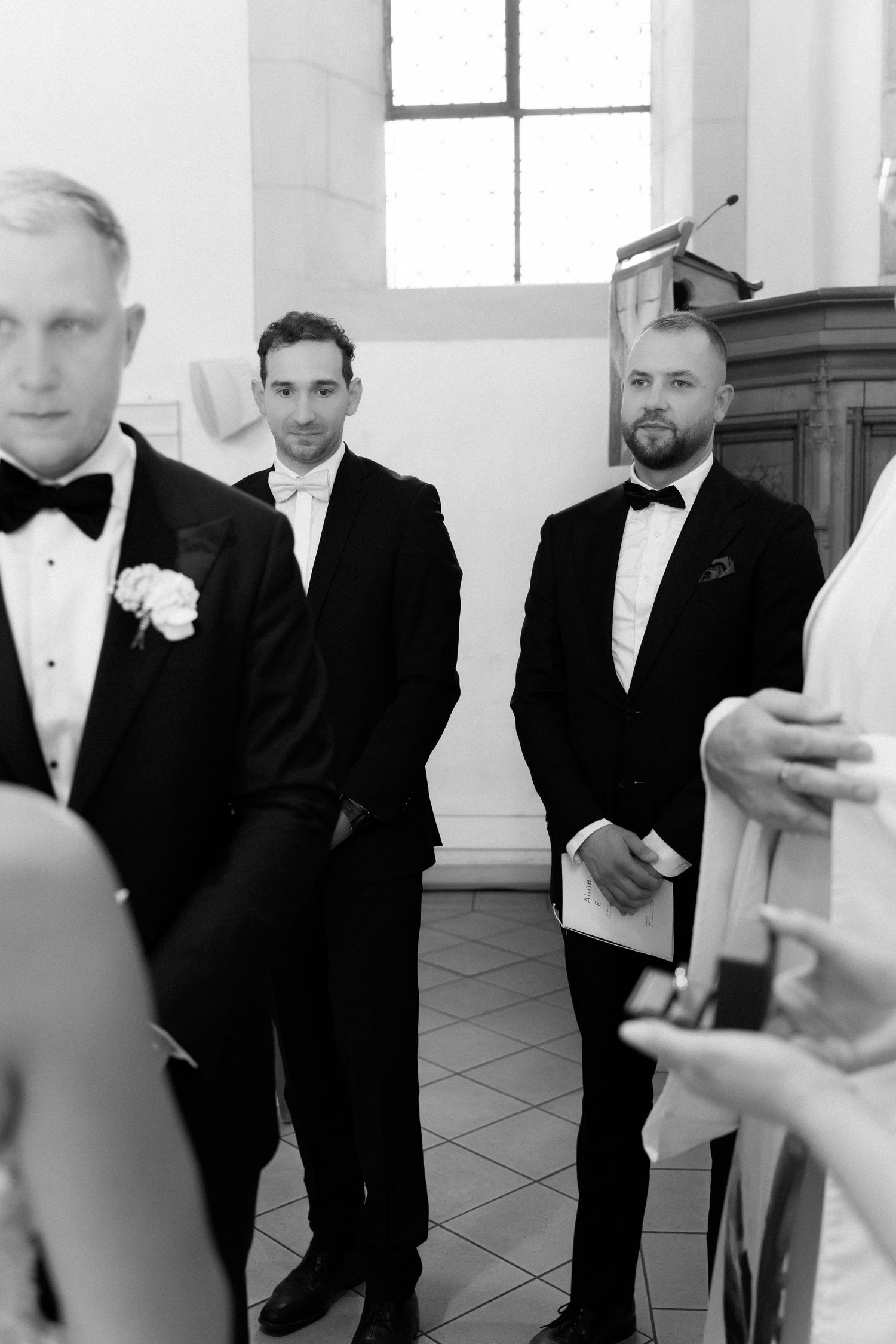 Three groomsmen in dark suits and bow ties stand and watch during a wedding ceremony in a bright church, in black and white.